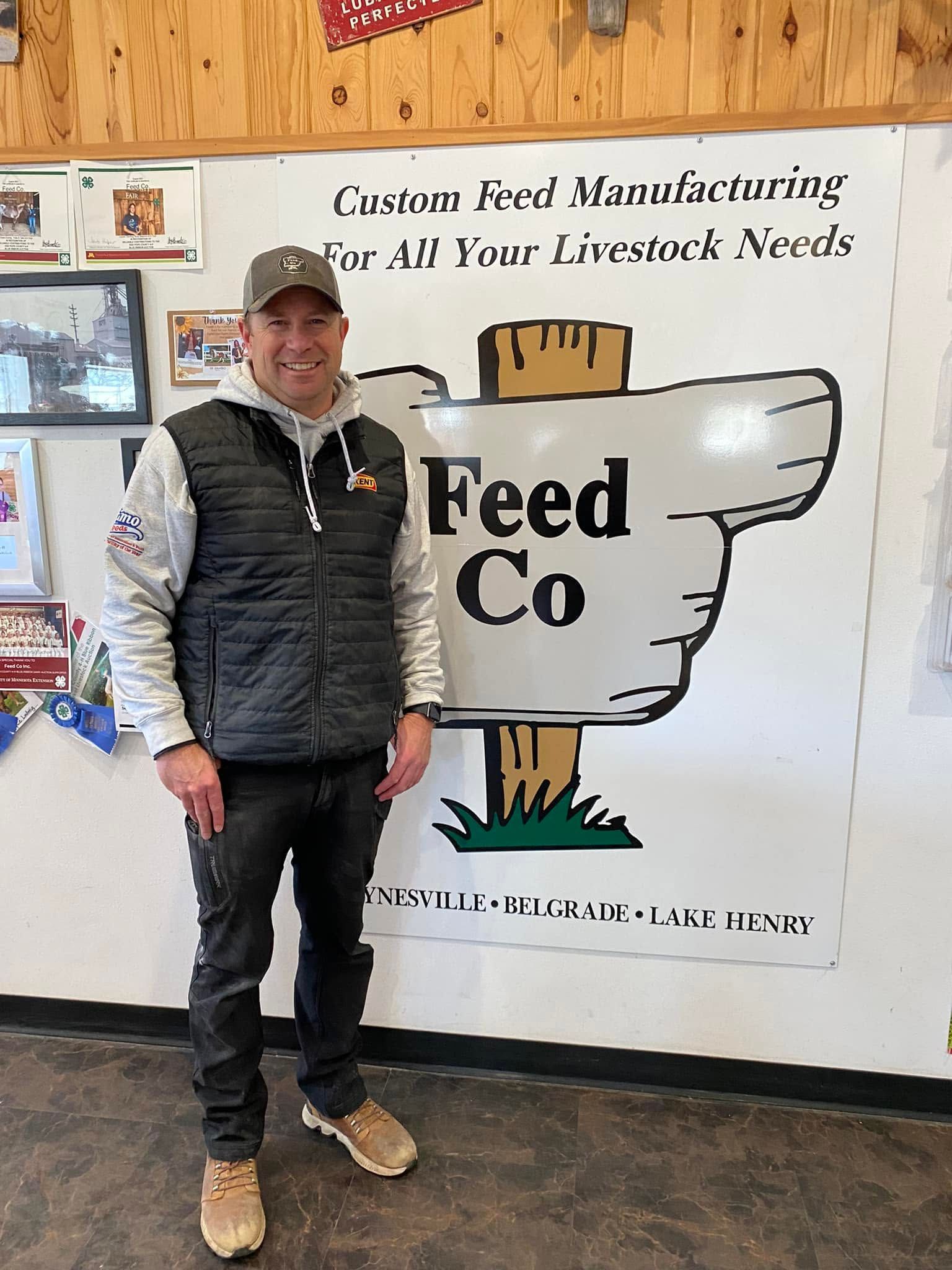 Man standing beside a Feed Co mural in a rustic shop interior, wearing a cap, vest, jeans, and work boots