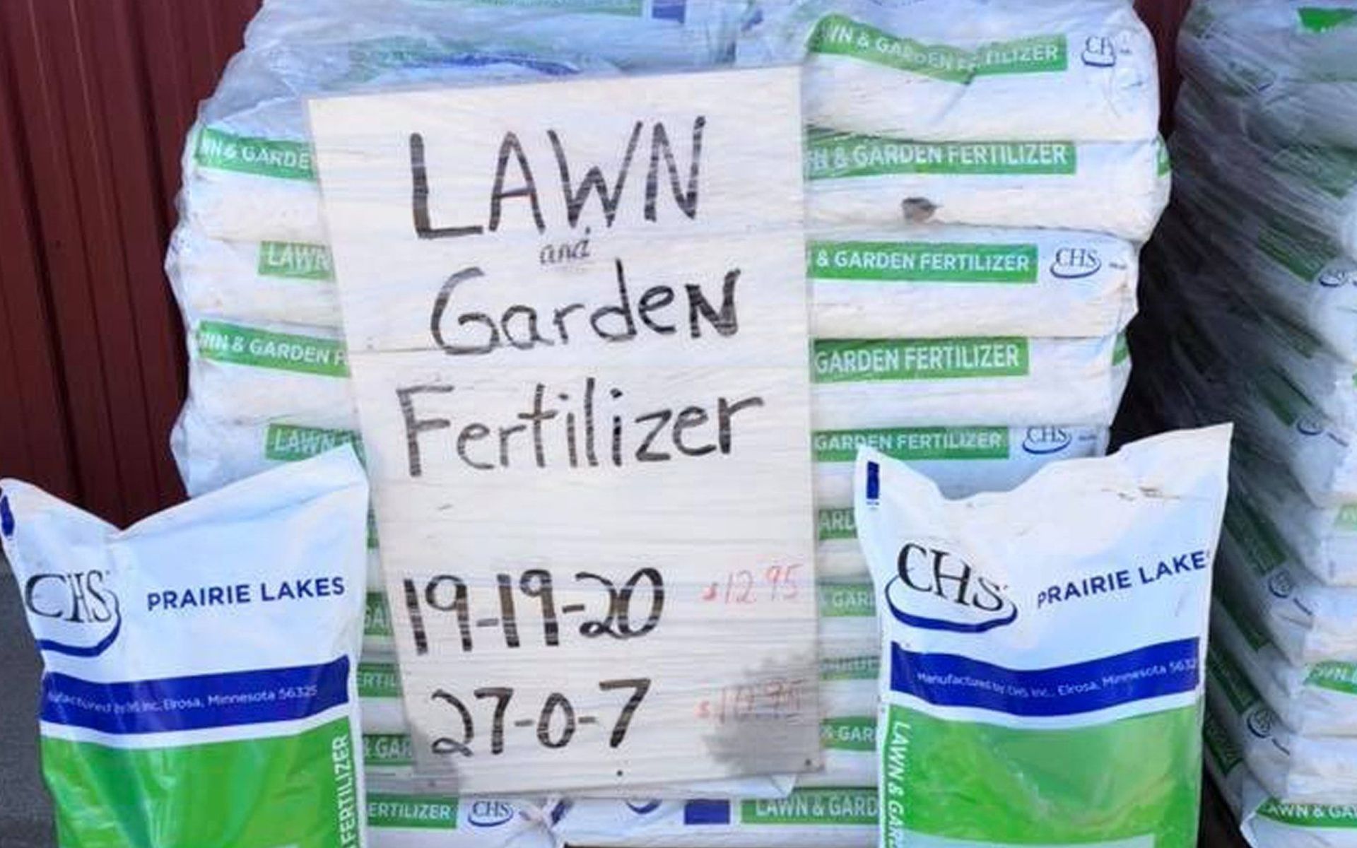 Stacked lawn and garden fertilizer bags with a handwritten sign listing numbers and dates.