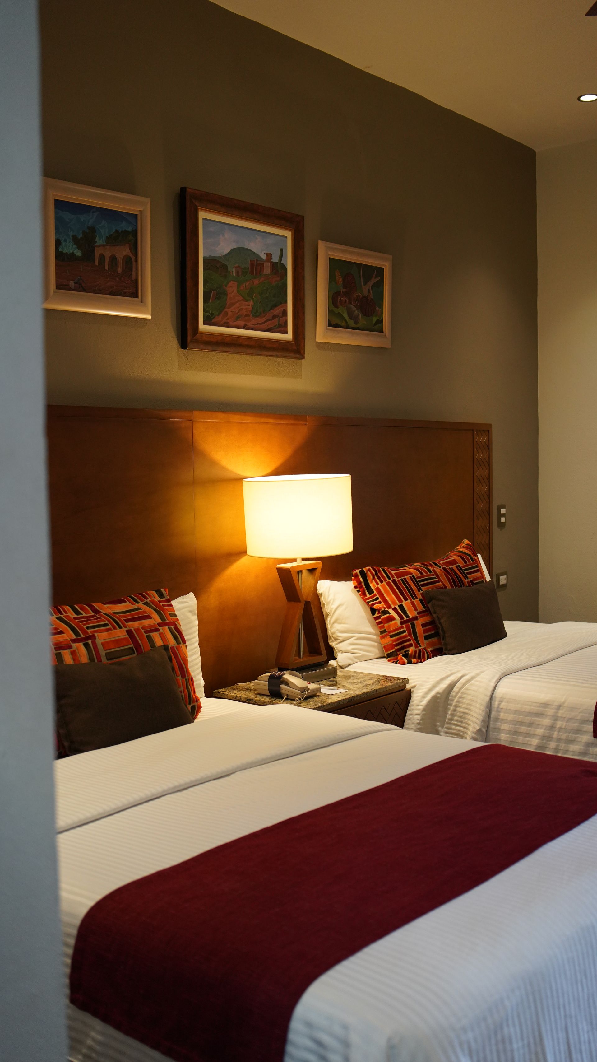 A hotel room with two beds , a lamp and paintings on the wall.