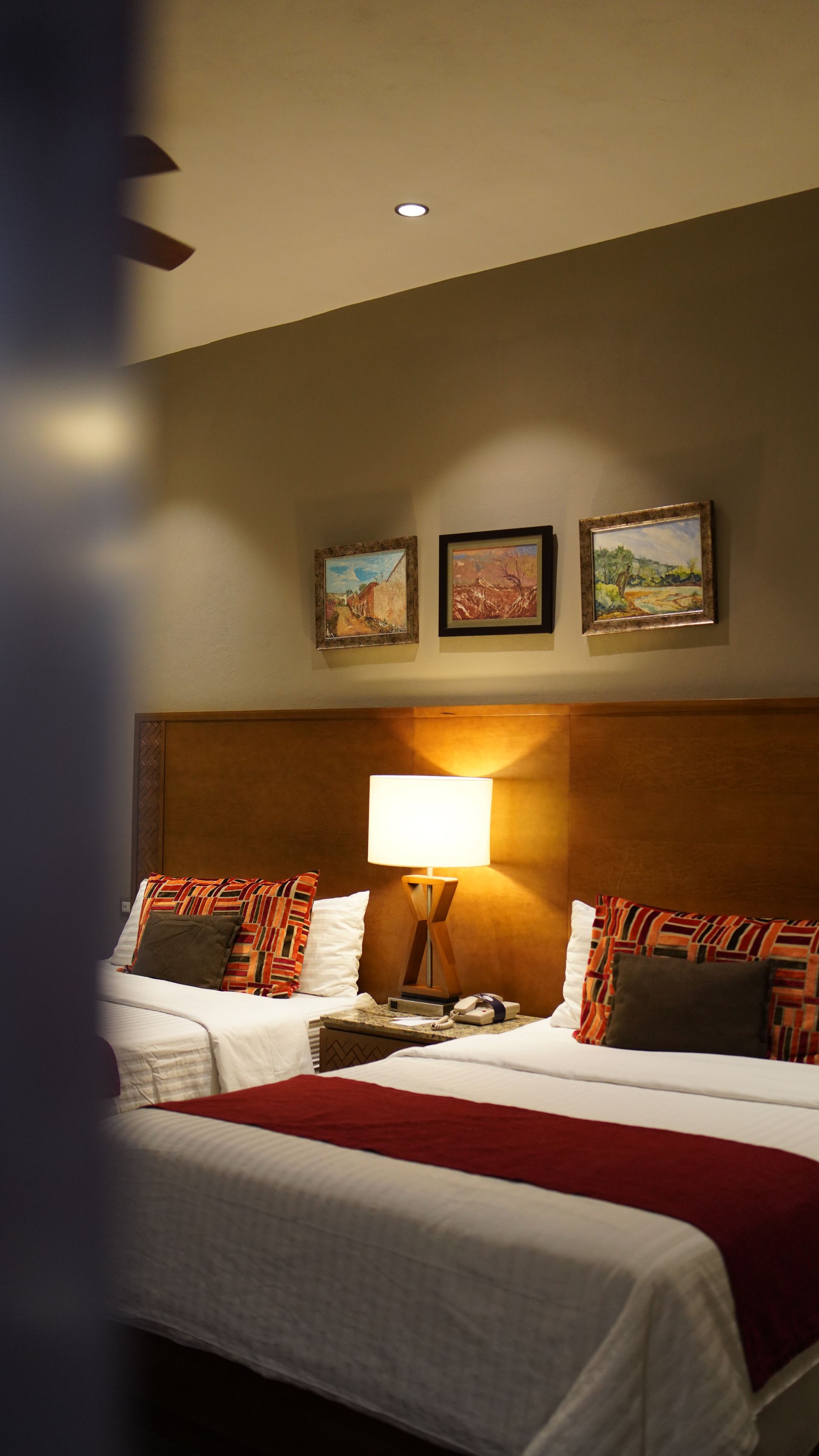 A hotel room with two beds , a lamp , and paintings on the wall.