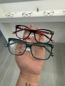 Una mano sostiene dos monturas de gafas, una roja y otra verde oscuro, delante de un estante lleno de gafas.