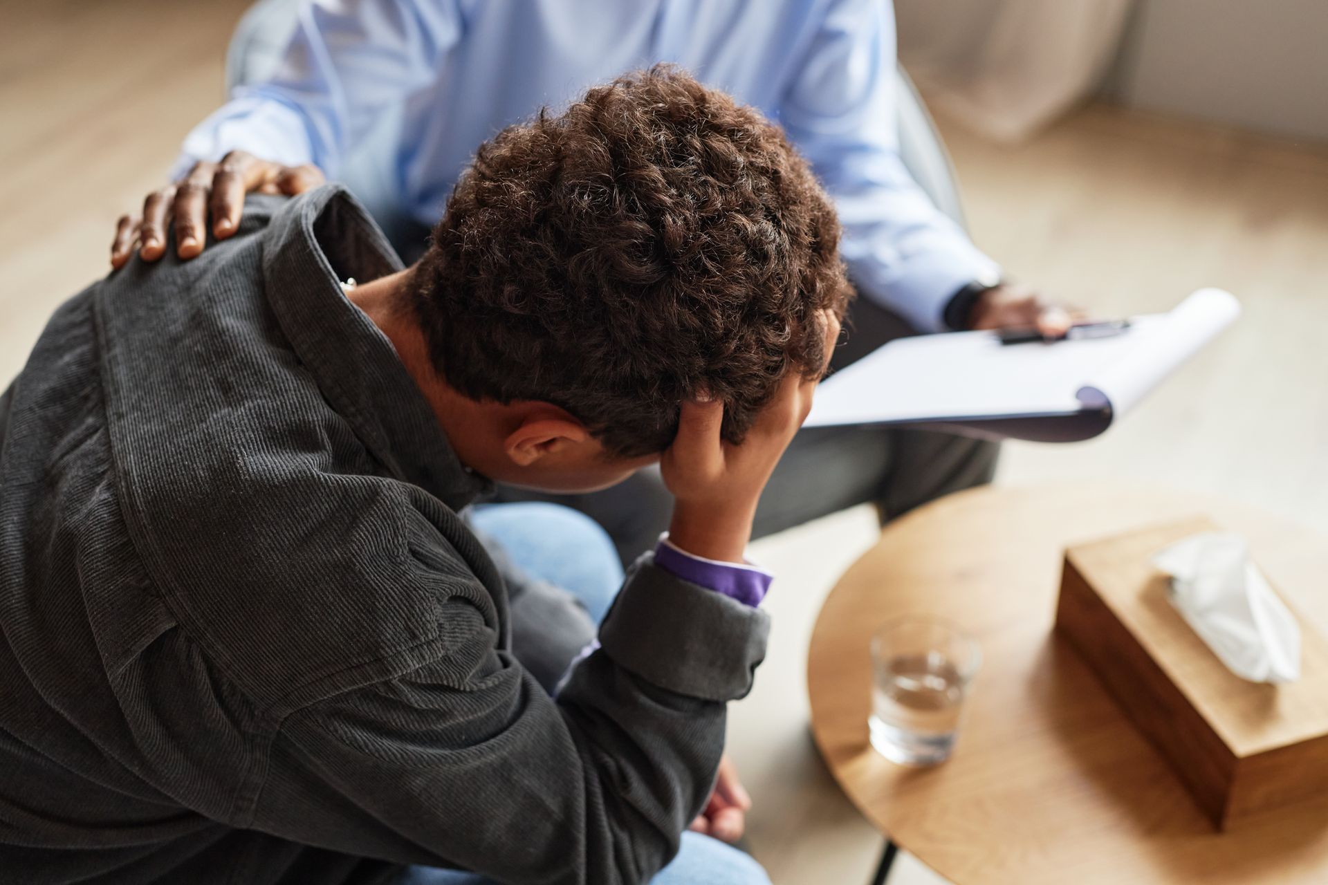 Person sitting with head in hand during a counseling session, with a therapist nearby and papers on a table