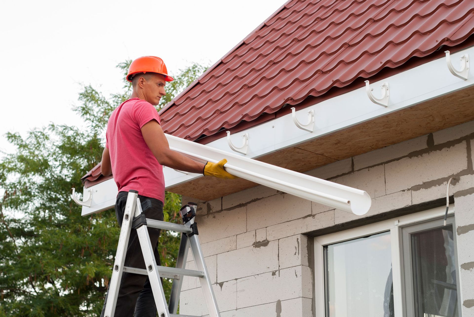 A man is standing on a ladder installing a gutter on the side of a house.