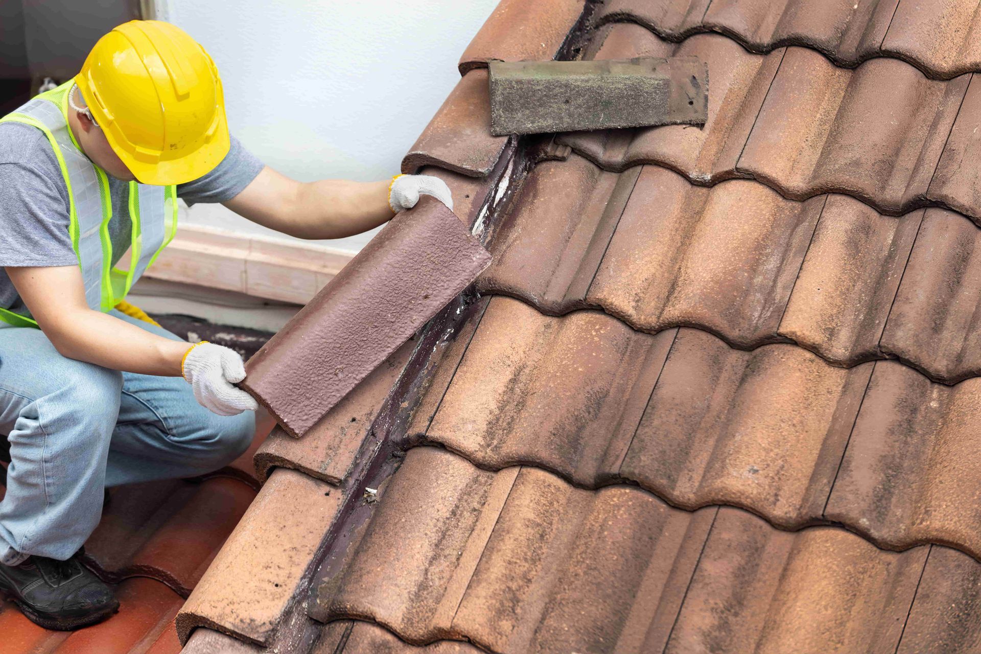 Worker repairing a brown tiled roof with a trowel and mortar, wearing a yellow hard hat and safety vest