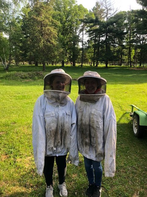 Two smiling girls in beekeeping suits standing in a sunny, grassy area surrounded by trees.