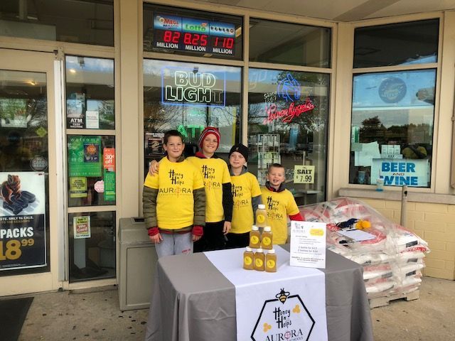 Four kids in yellow Honey Helps shirts stand behind a table with jars of honey outside a store, set up for a fundraising event.