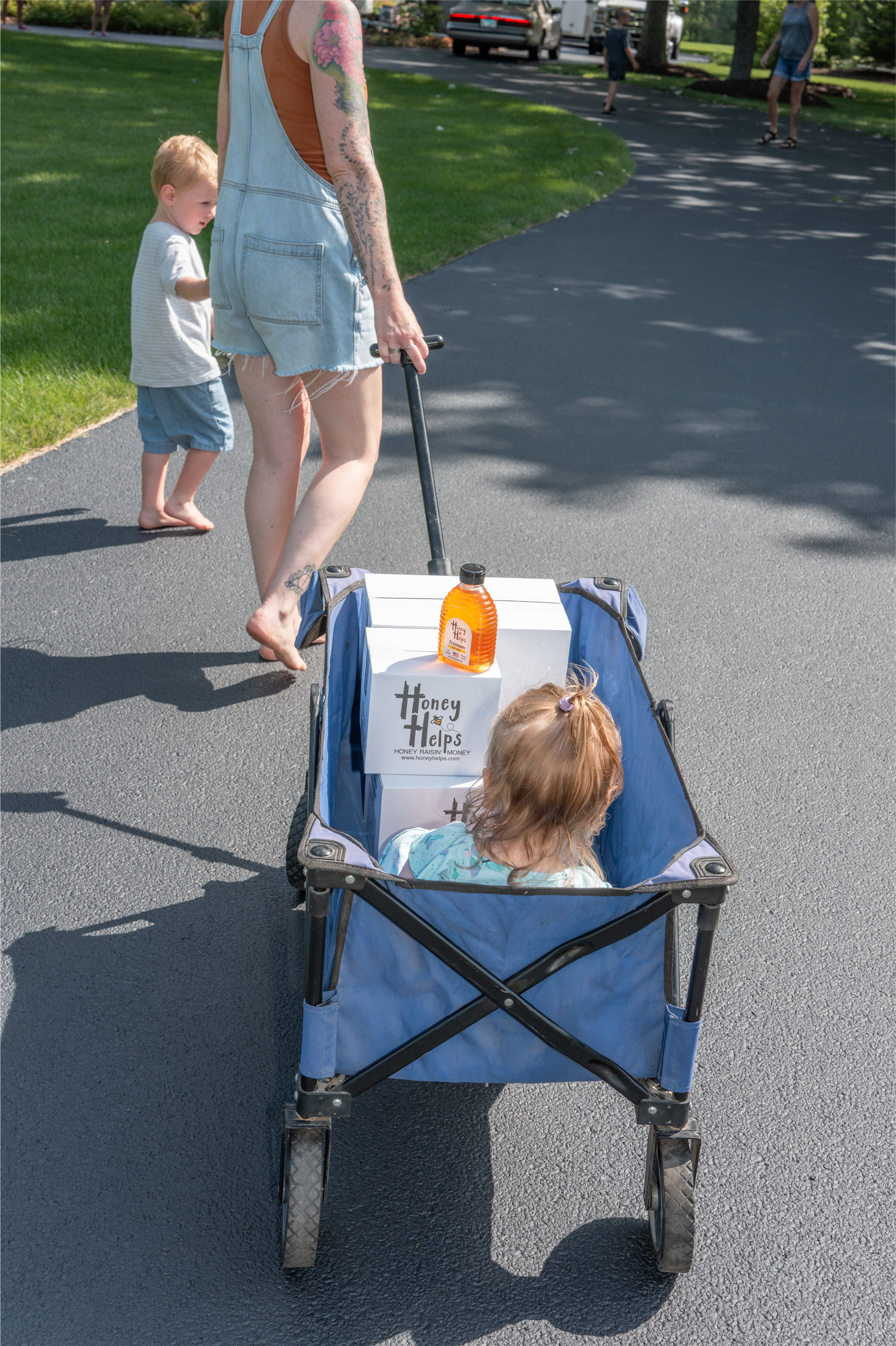 Blue wagon with a child inside being pulled along a paved park path by an adult with a toddler nearby