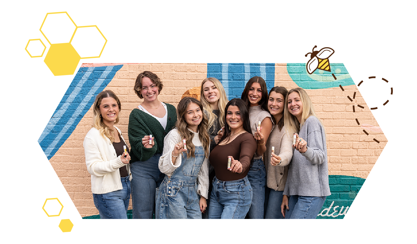 Group of women holding Honey Helps lip balm, smiling together in front of a colorful mural
