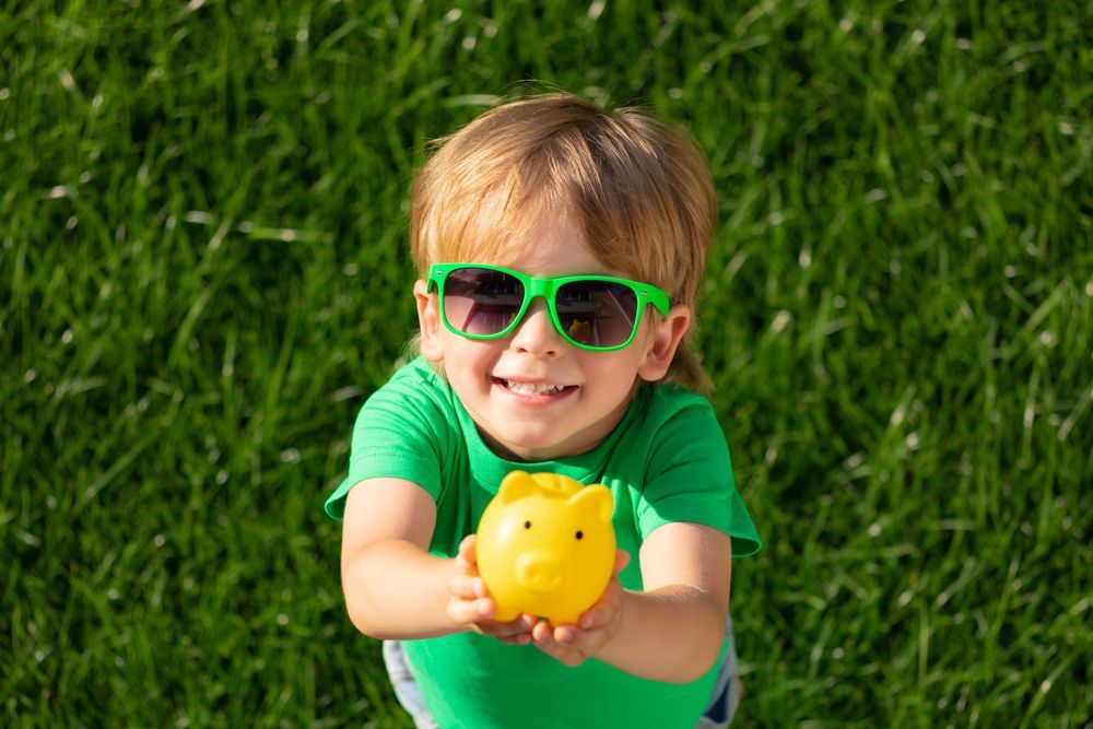 Child holding piggy bank in hands against green spring background.