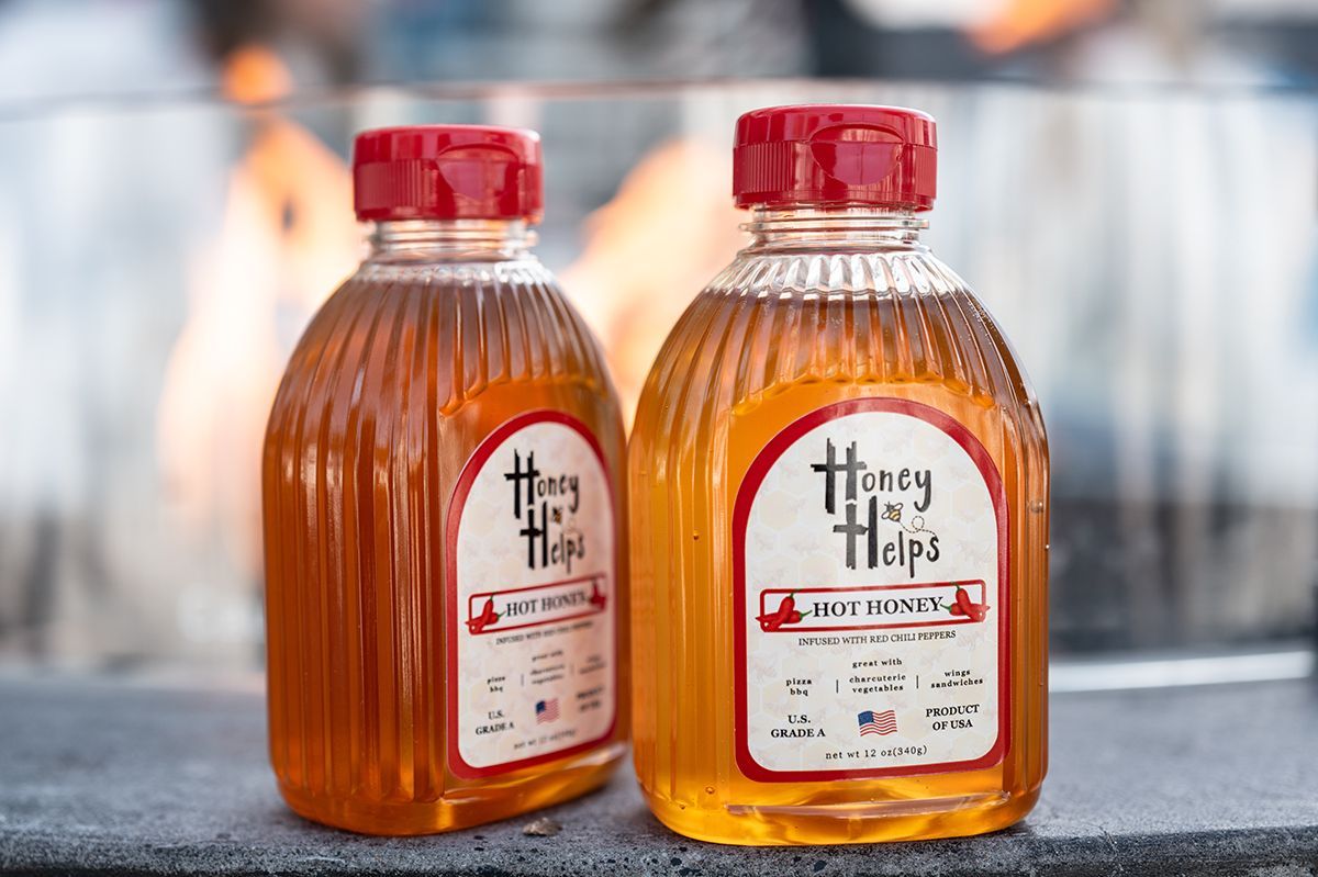 Two bottles of Honey Helps hot honey infused with red chili peppers on outdoor table with fire background
