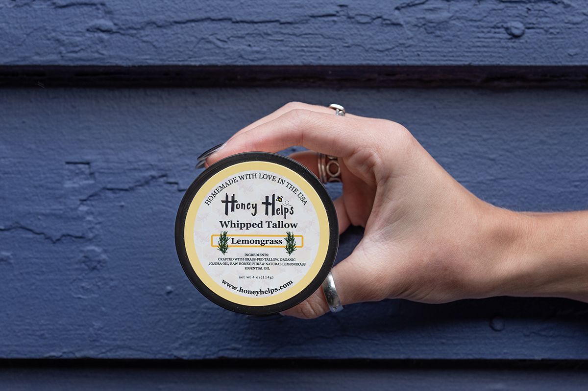 Hand holding Honey Helps whipped tallow lemongrass balm against blue textured wall background