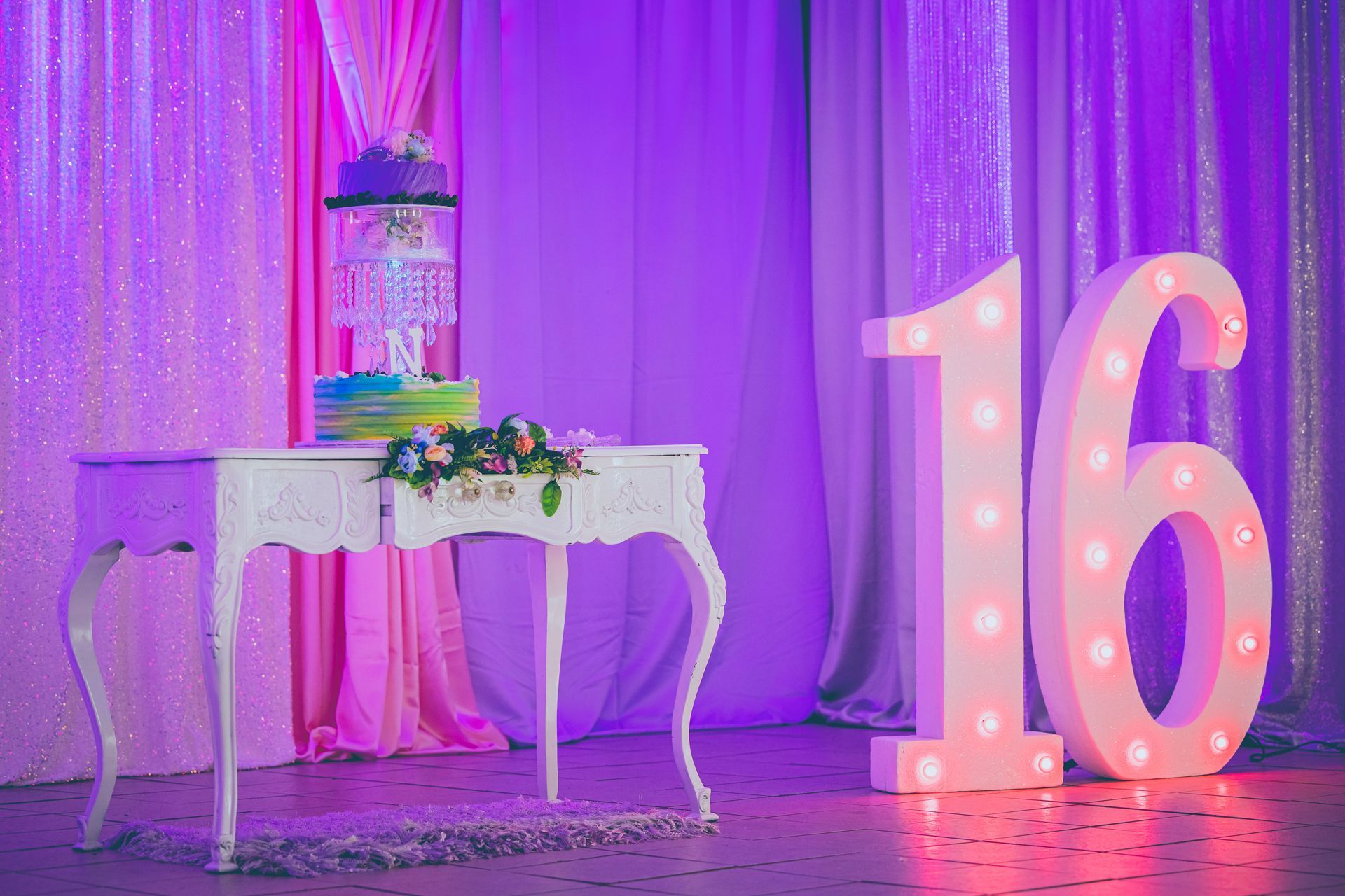 Purple-lit party setup with a decorated table, cake, and large illuminated number 16 sign