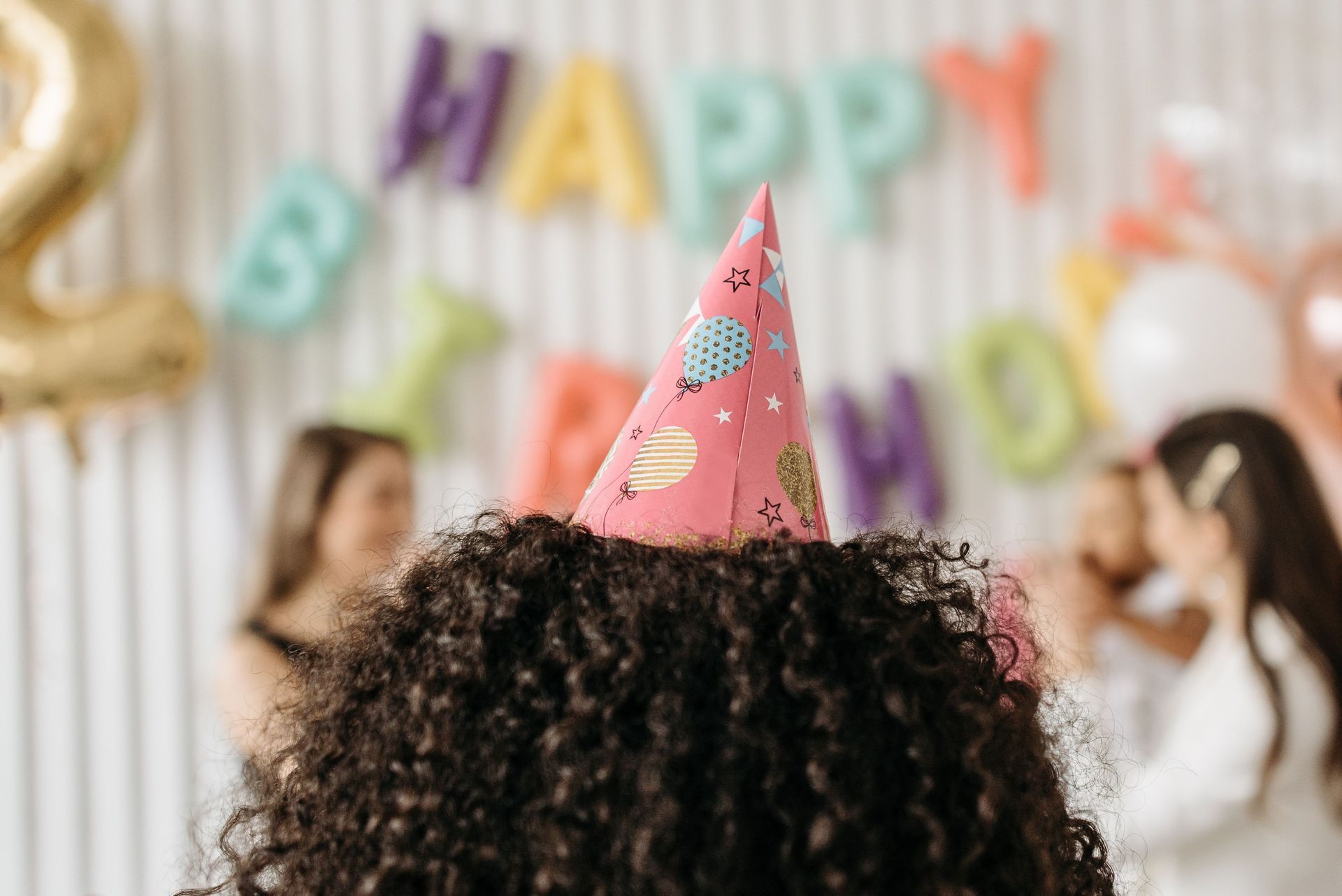 Party guests wearing colorful cone hats at a birthday celebration with a HAPPY BIRTHDAY banner