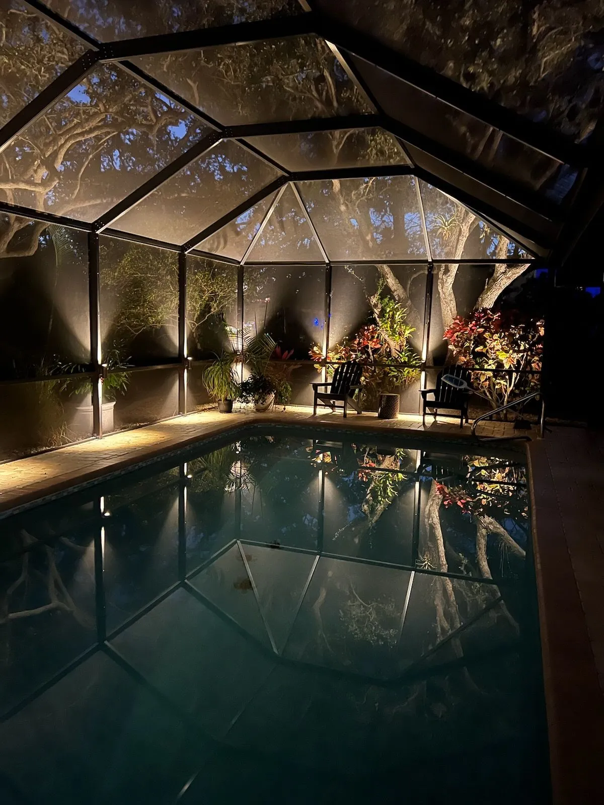 Indoor pool at night under a glass enclosure, lit by warm string lights and surrounded by plants.