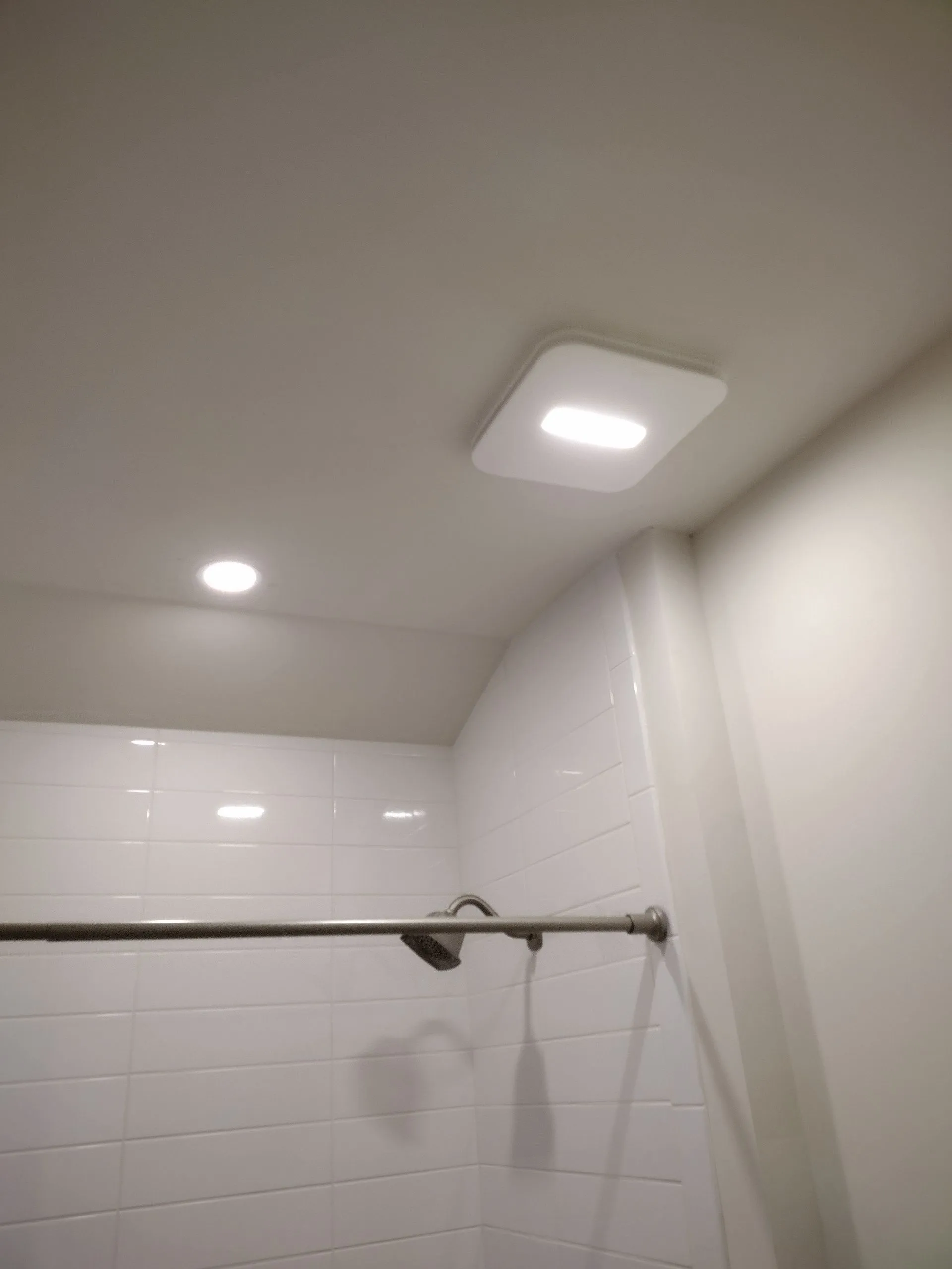 Bathroom shower with white tiled walls, ceiling lights, and a chrome shower rod