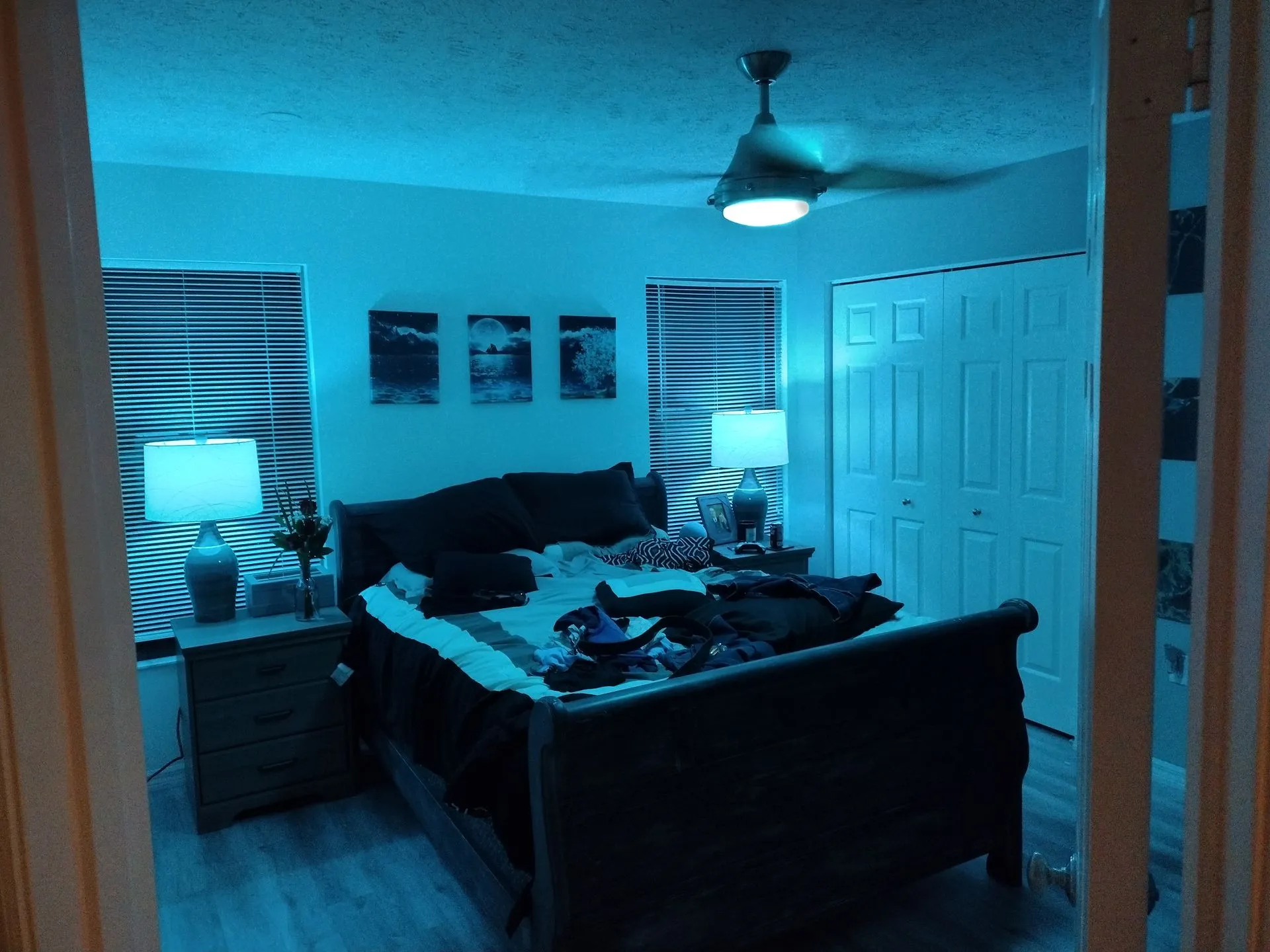 Blue-lit bedroom with unmade bed, ceiling fan, dresser lamp, wall art, and closet doors