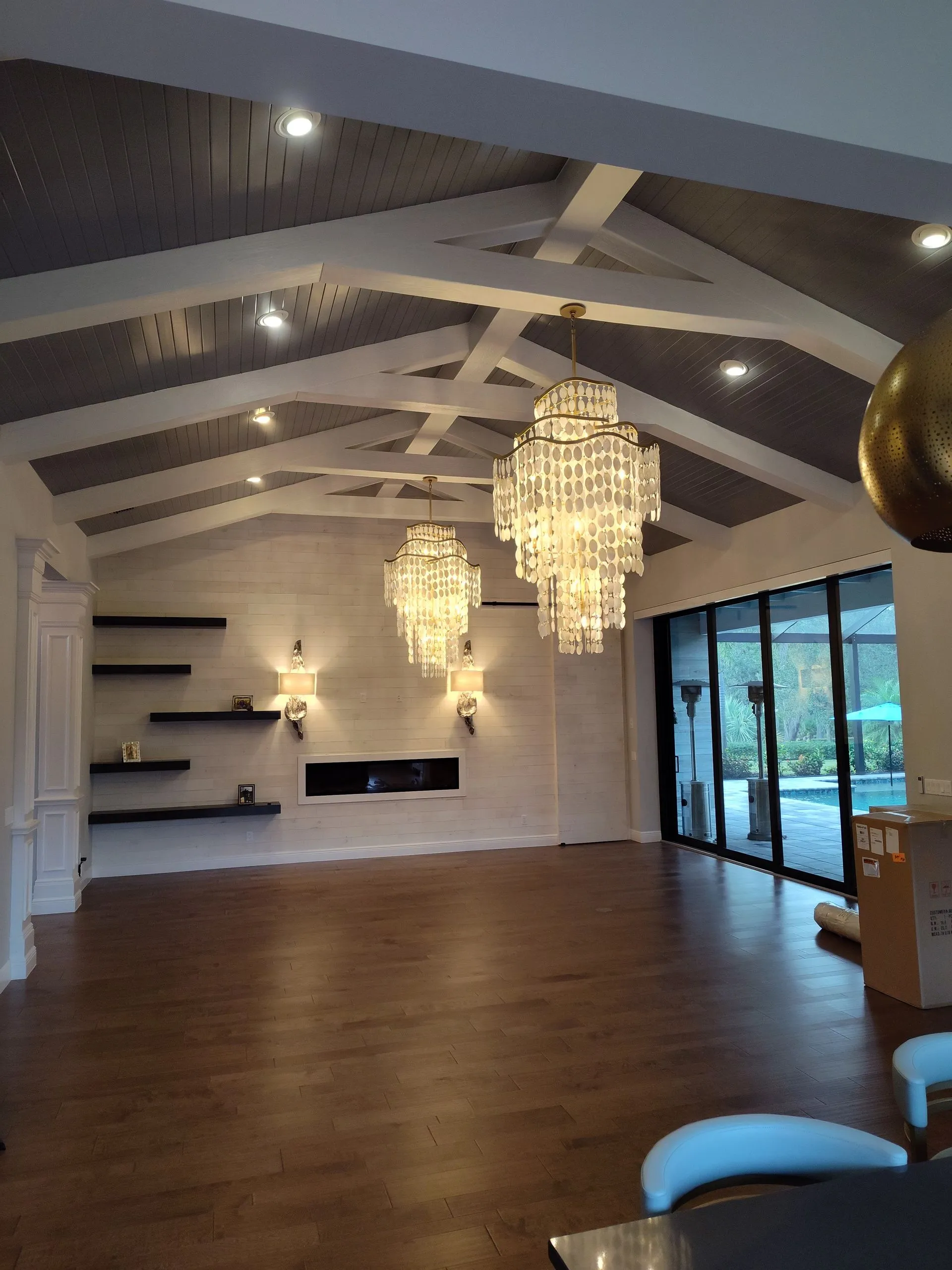Spacious modern room with wood floors, white beams, chandeliers, shelves, and large glass doors.