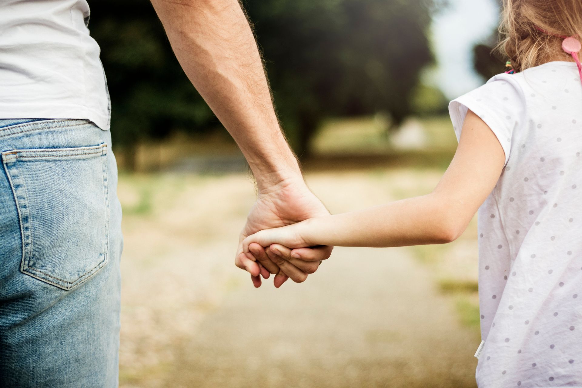 Person in jeans holds a child's hand while walking on a path, with greenery in the background.