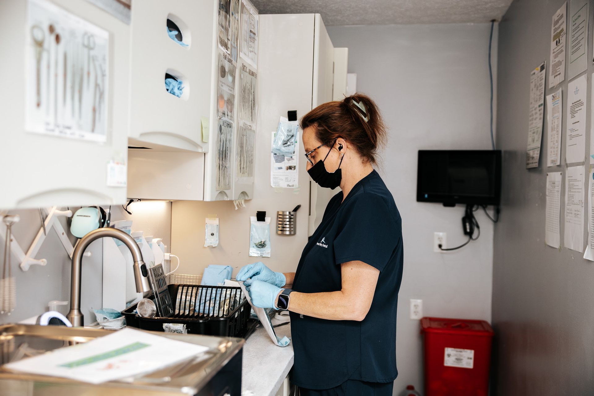 Dentist in scrubs and mask prepares instruments at a sink in a dental office.