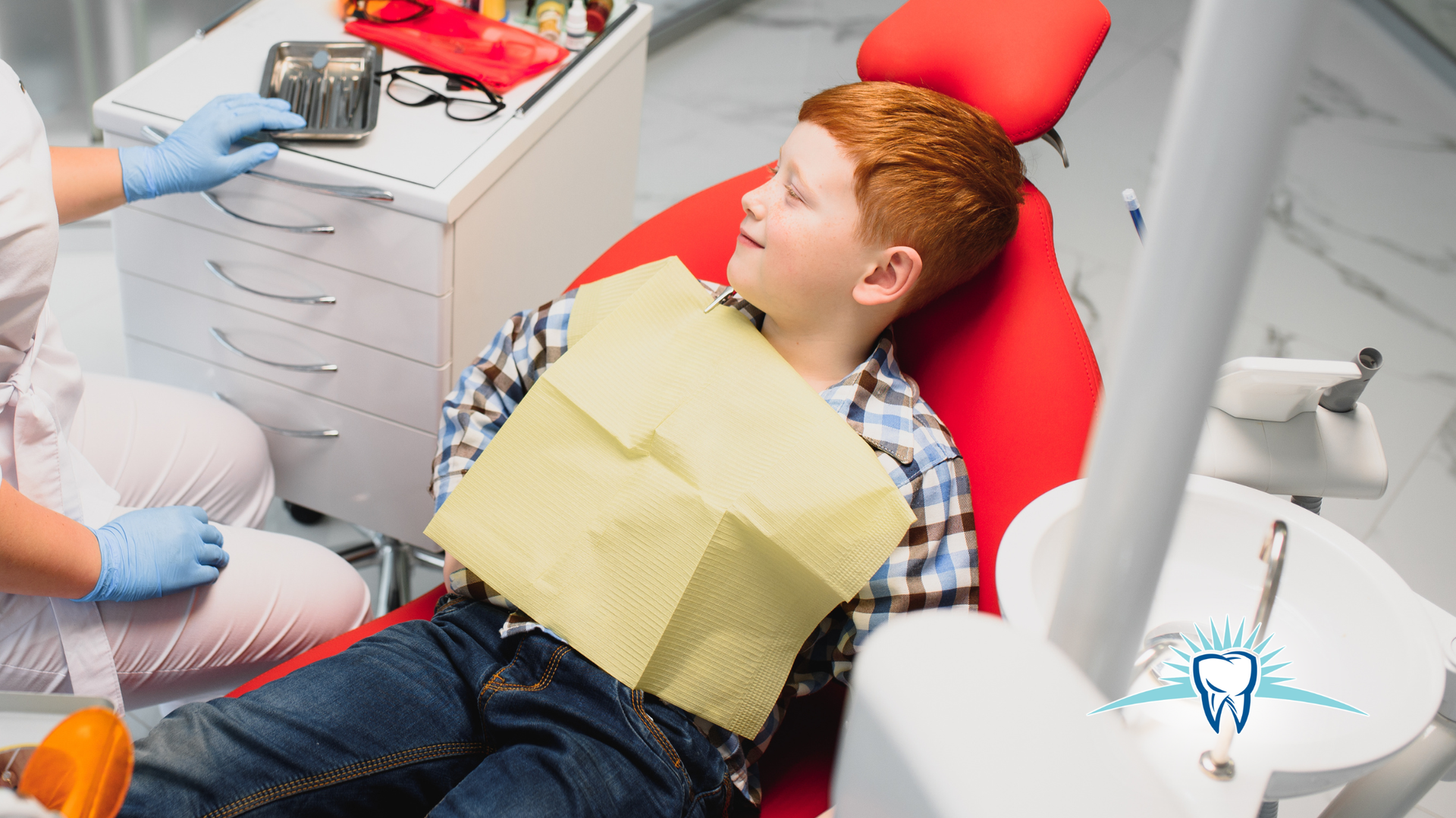 Boy in dental chair with bib, looking at a dental assistant. Clinic setting.