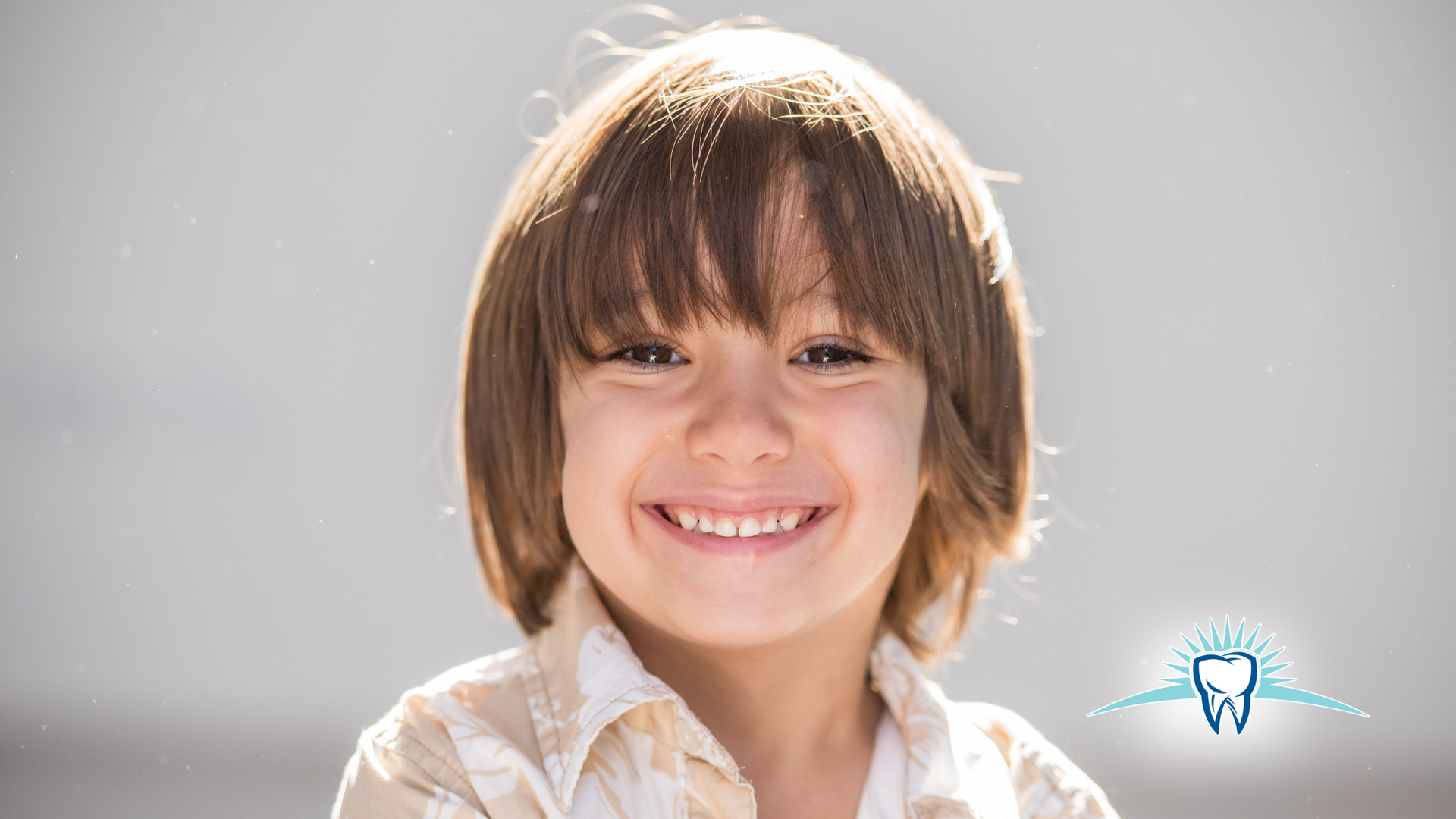 Smiling child with long brown hair, wearing a light-colored shirt, against a bright background.