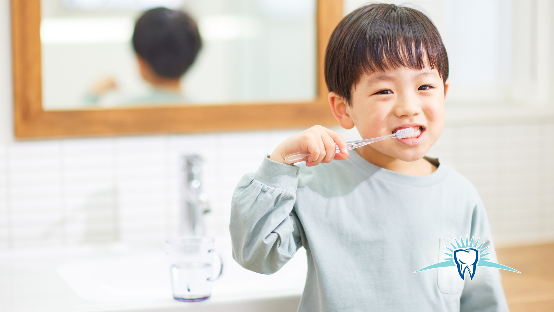 Boy brushing teeth in a bathroom, smiling. He holds a toothbrush in front of a mirror and sink.