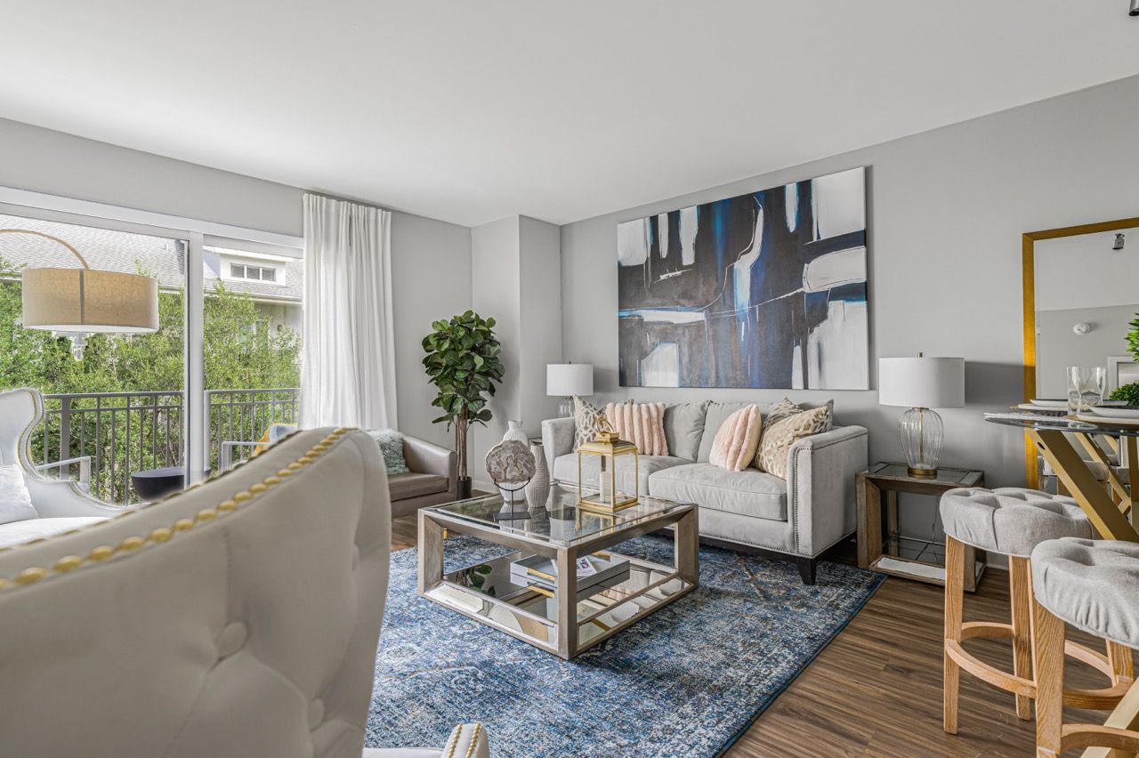 Bright living room in a modern apartment with a gray sofa, glass coffee table, artwork, and balcony doors.