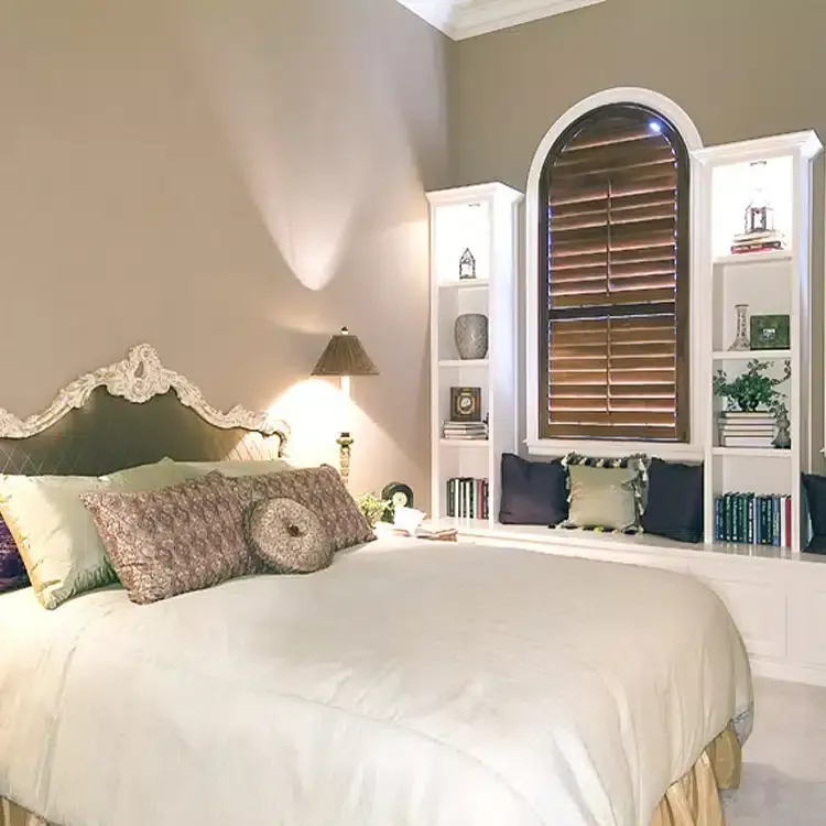 A cozy bedroom with a bed, bookshelves, and a window adorned with wood plantation shutters.