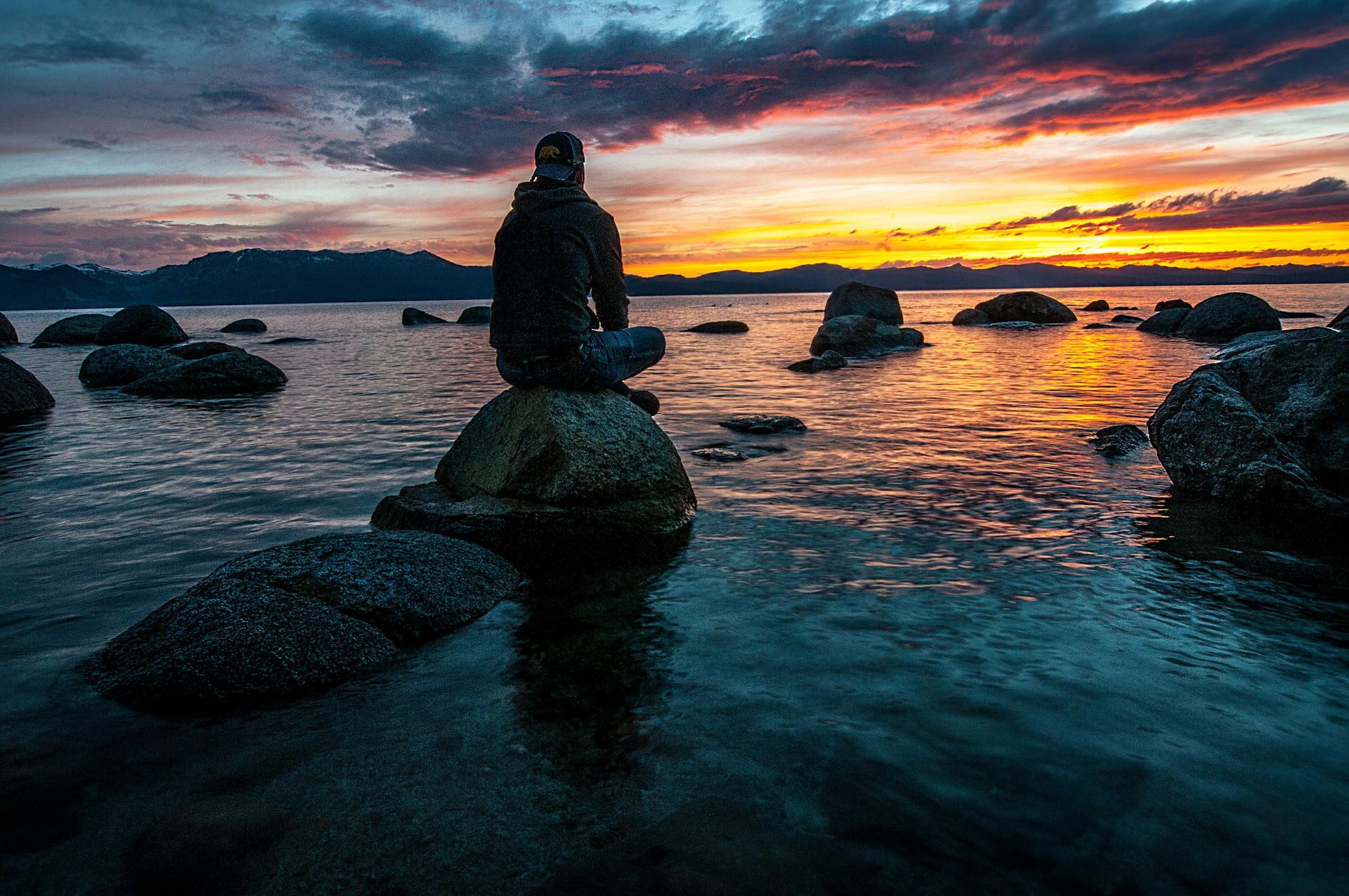 Person sitting on rock, facing sunset over water. Orange and yellow sky, dark water and rocks.