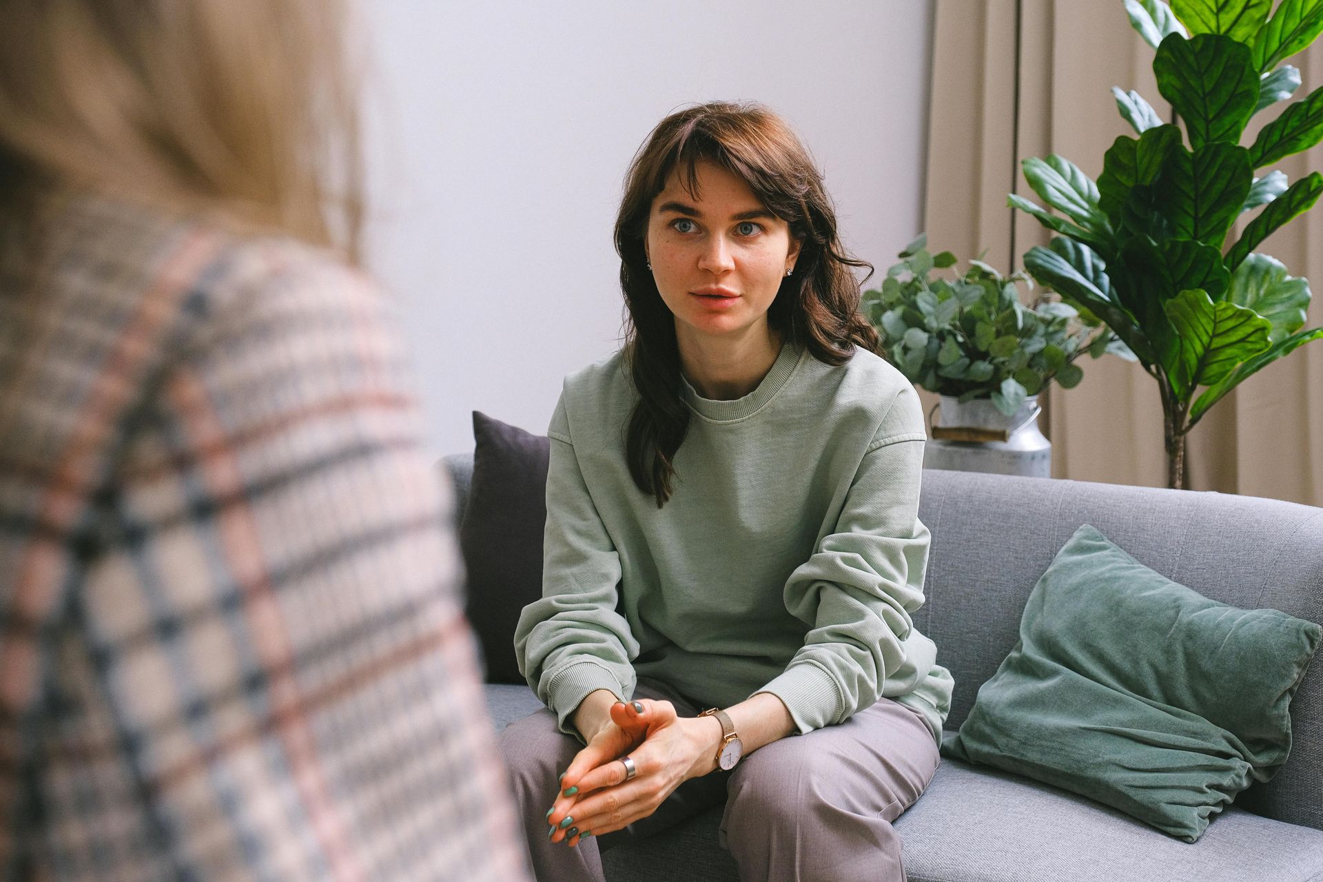Two people sit on a couch during a conversation, with a potted plant nearby in a brightly lit indoor space.