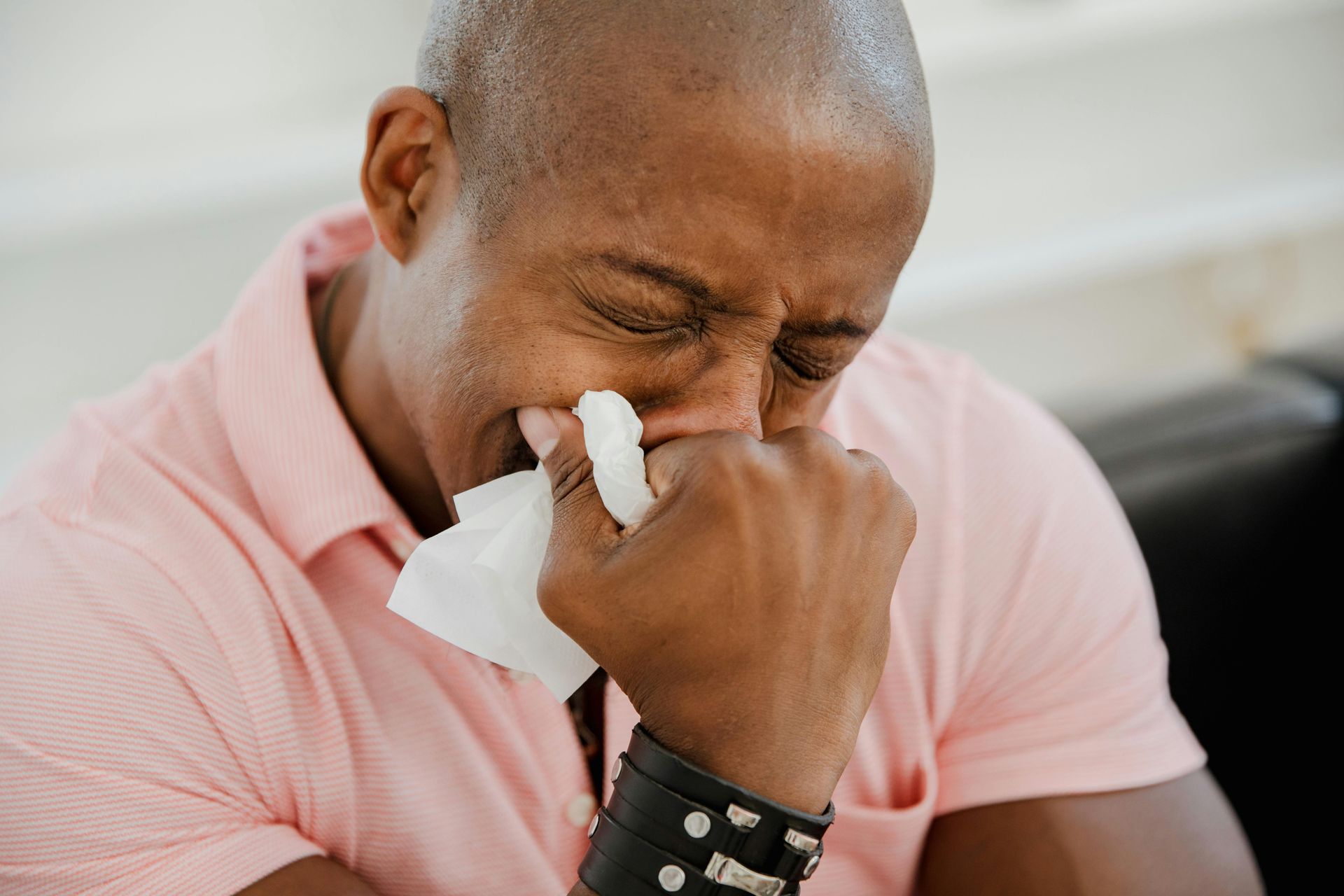 Man in pink shirt, wiping nose with tissue, eyes closed in a seated indoor setting.