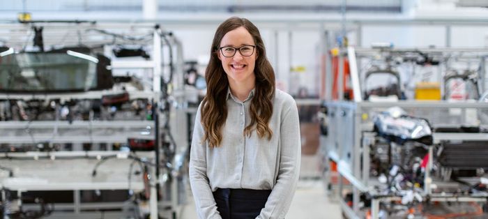 A woman is standing in a factory with a car in the background.