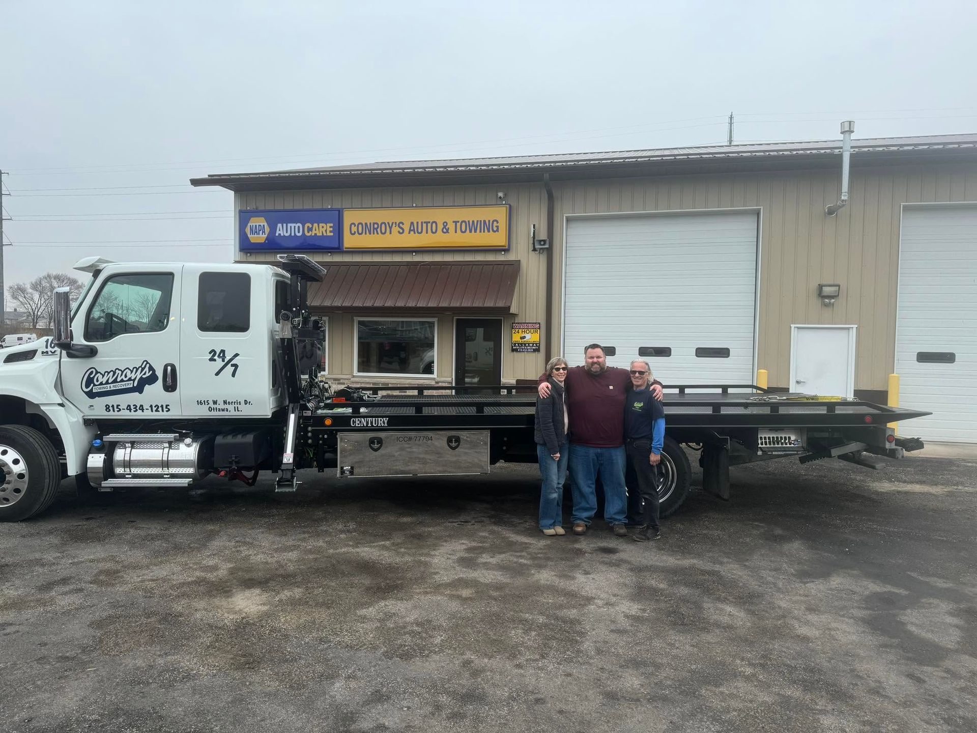 White utility truck parked by a warehouse with three workers standing beside it on a wet lot.