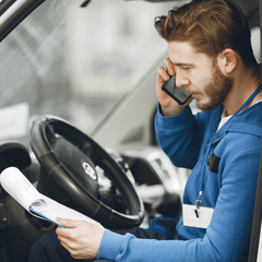 A man in a blue hoodie sits in a vehicle, reading paperwork while on a phone call.