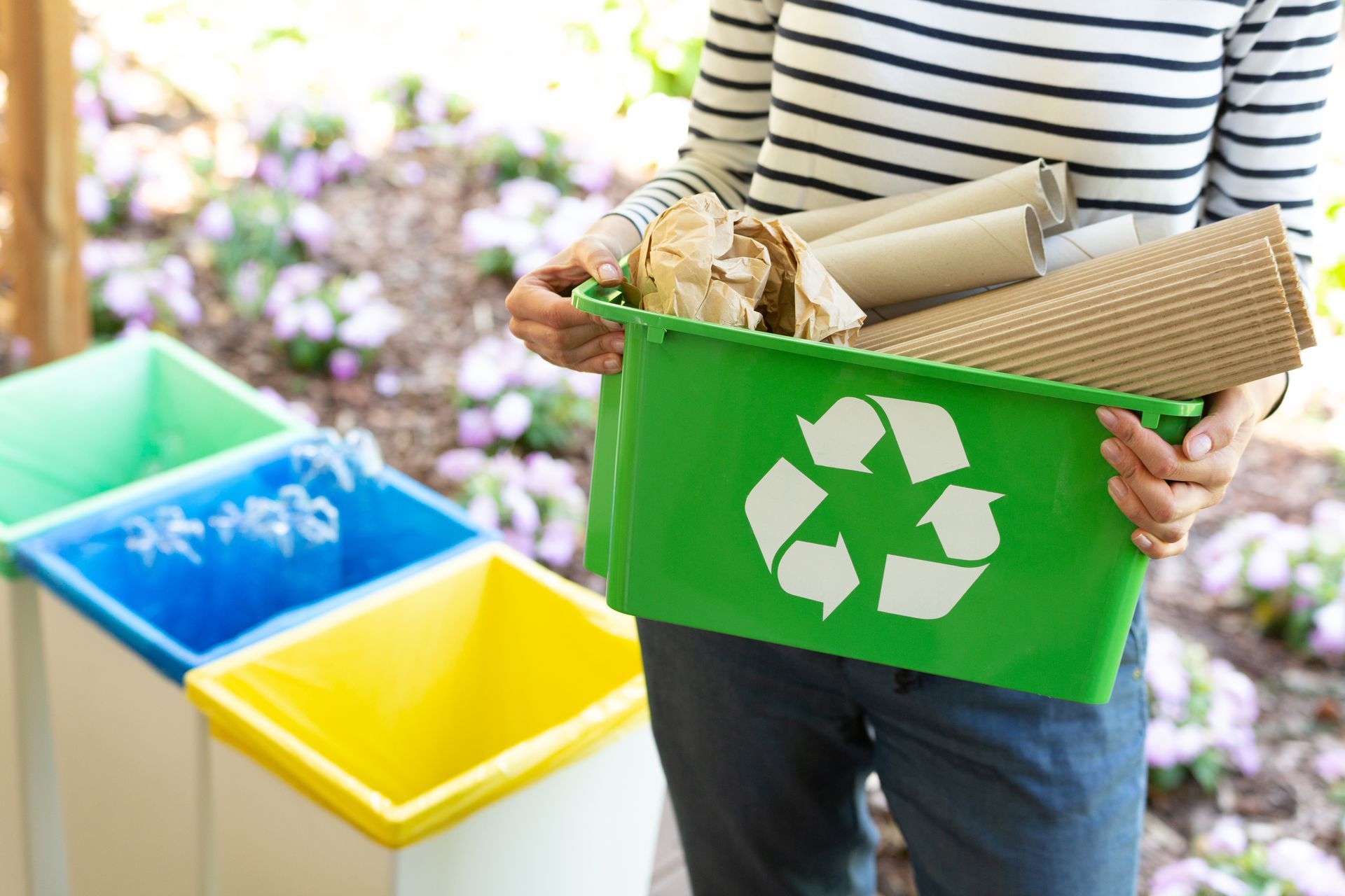 Person holding green recycling bin filled with paper near sorting containers outdoors. Person holding green recycling bin filled with paper near sorting containers outdoors.