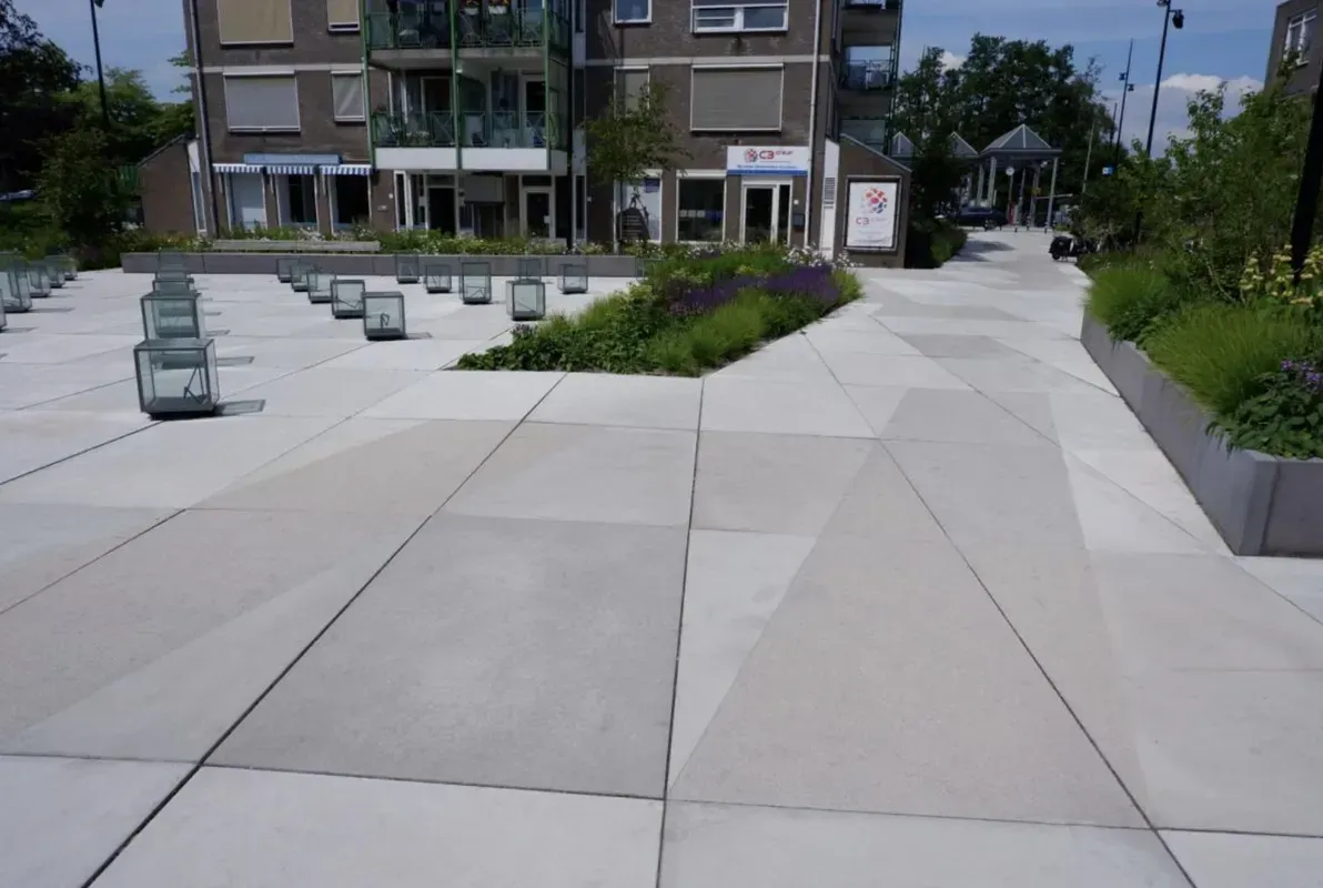 A modern public square with large concrete pavers, small metal lanterns, and landscaped planters in front of an apartment.