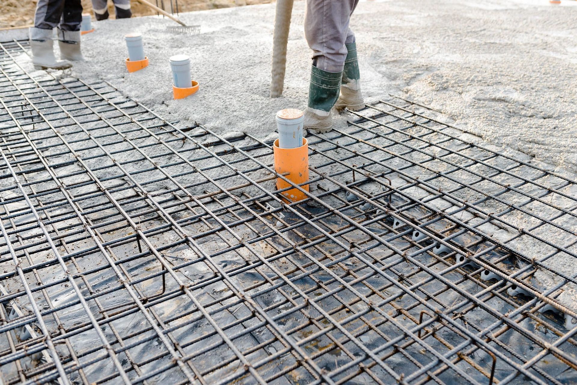 Workers pour concrete over a steel reinforcement grid containing upright plumbing pipes on a construction site.