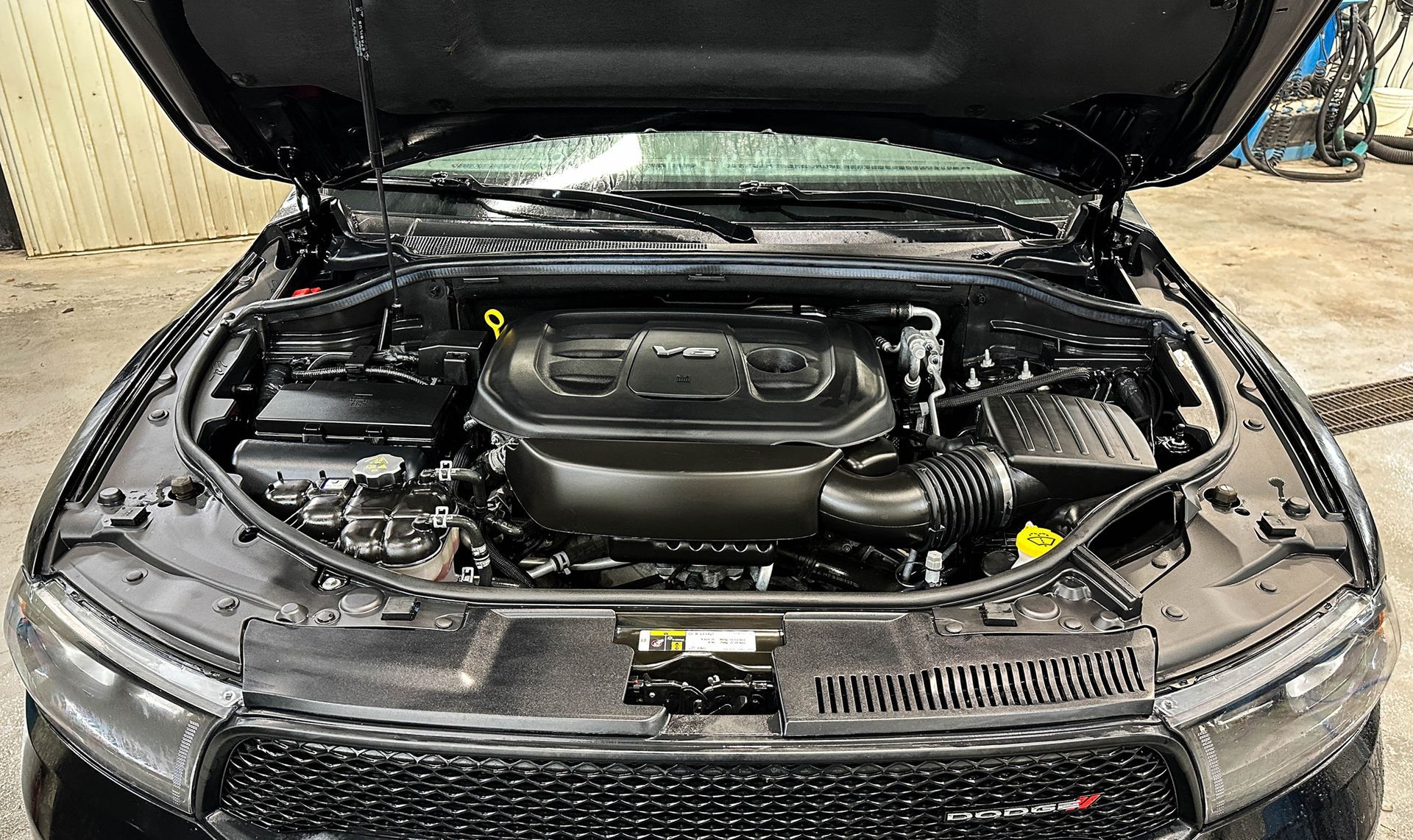 Open car hood showing engine bay and front grille, with a black vehicle in a garage.