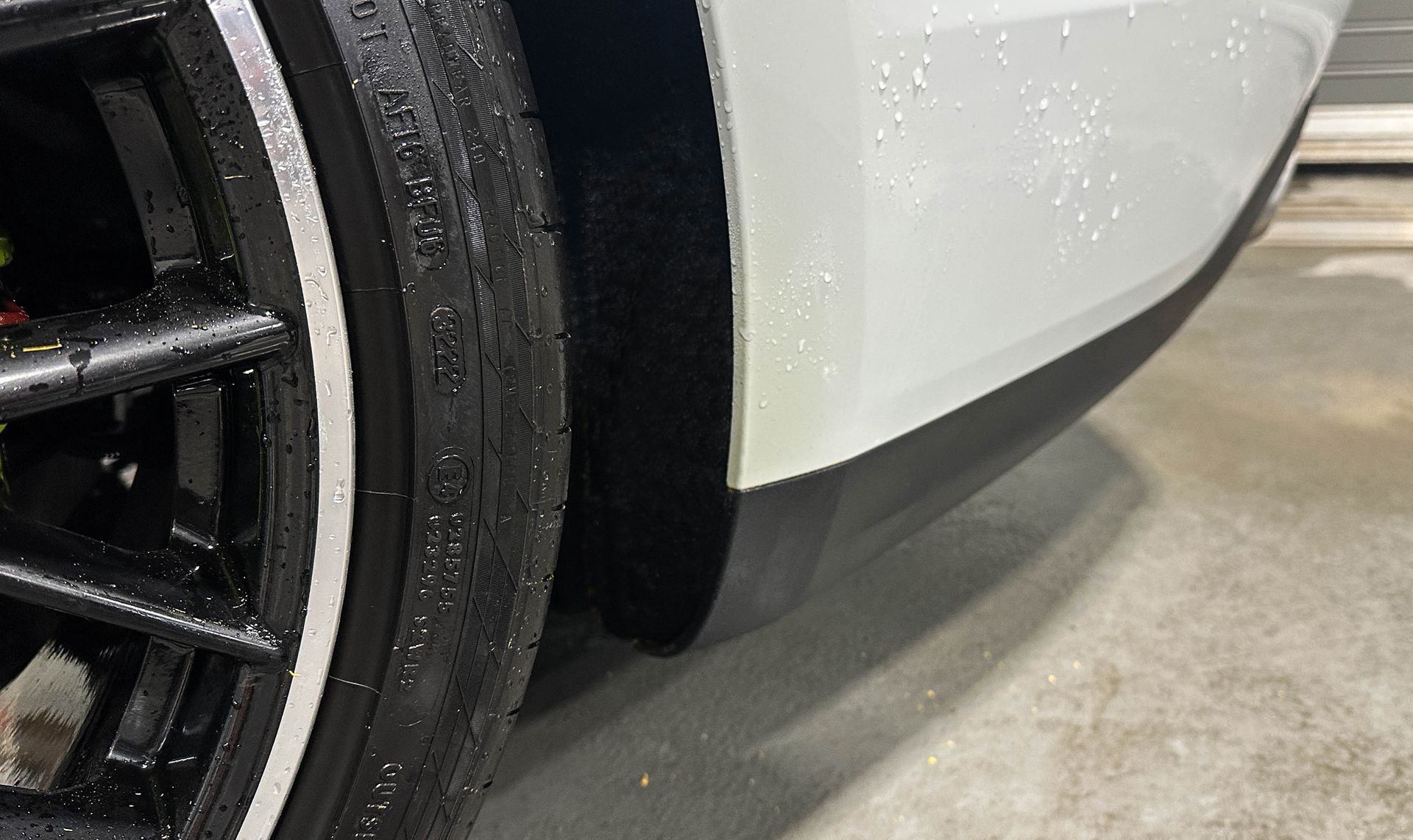 Close-up of a white car’s front wheel and bumper on a concrete floor, showing a black rim.