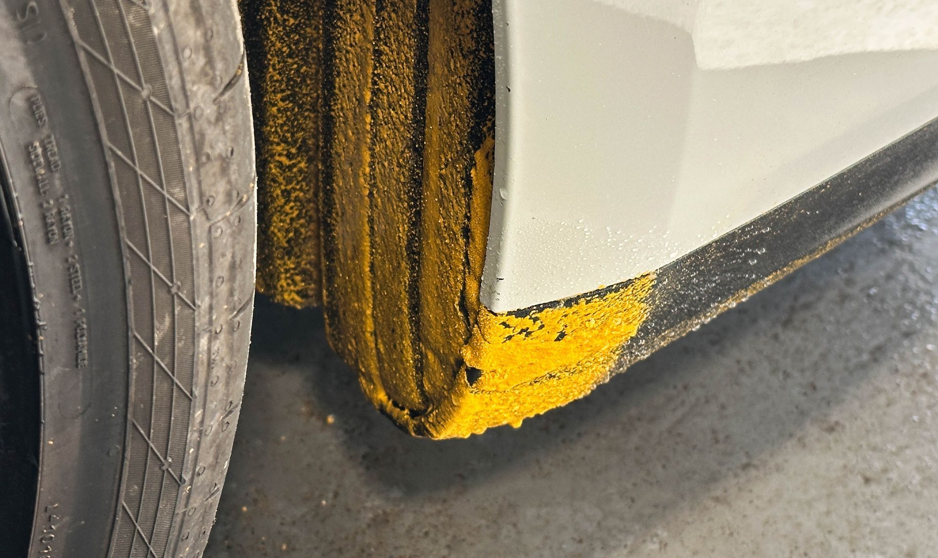 Yellow paint scrape on a white car’s wheel arch near the tire and lower bumper.