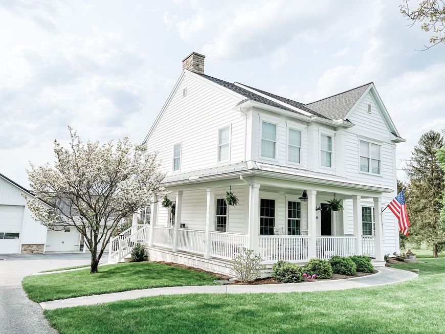 White two-story house with a front porch, blooming tree, and American flag on a green lawn