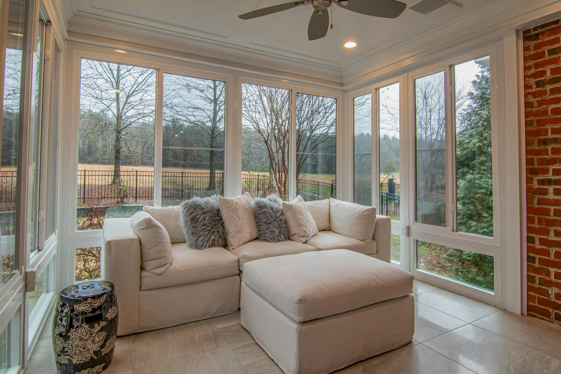 Bright sunroom with a beige sectional, ottoman, and large windows overlooking a yard.