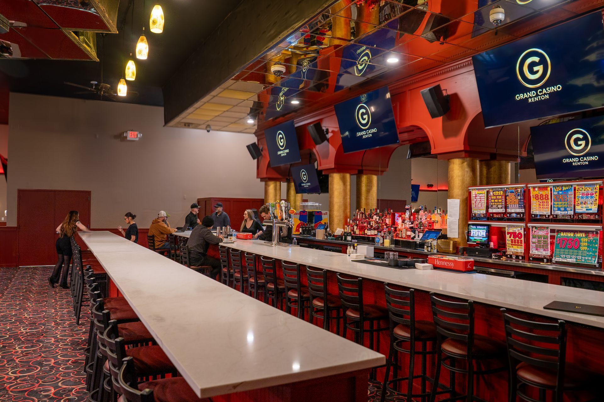 A row of tan bar stools lines a counter in front of a backlit wall logo and drink station at a sports bar.