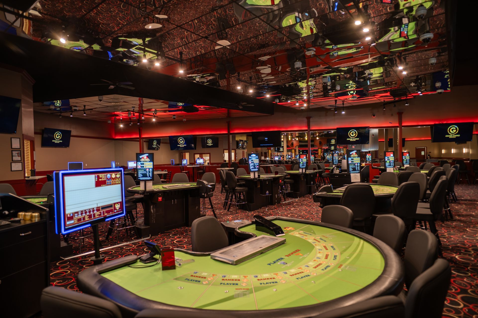 A modern, dim casino lounge featuring circular tables with chairs, screen monitors on walls, and patterned carpeting.