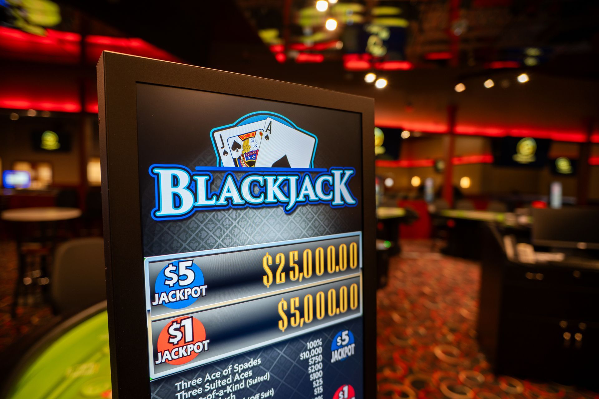 A dimly lit casino interior features rows of gaming tables with brown chairs and patterned carpeting under ornate lighting.