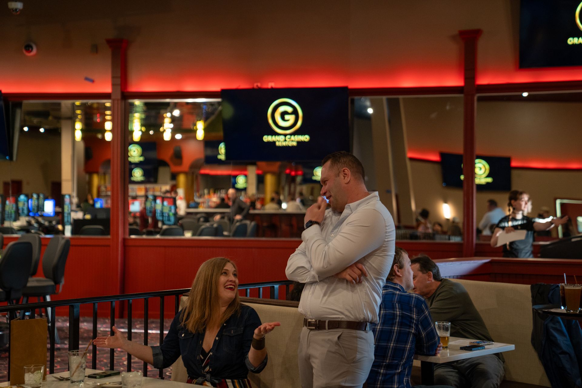 A casino floor featuring yellow game tables, rows of chairs, and a bar area with multiple TVs displaying sports programs.