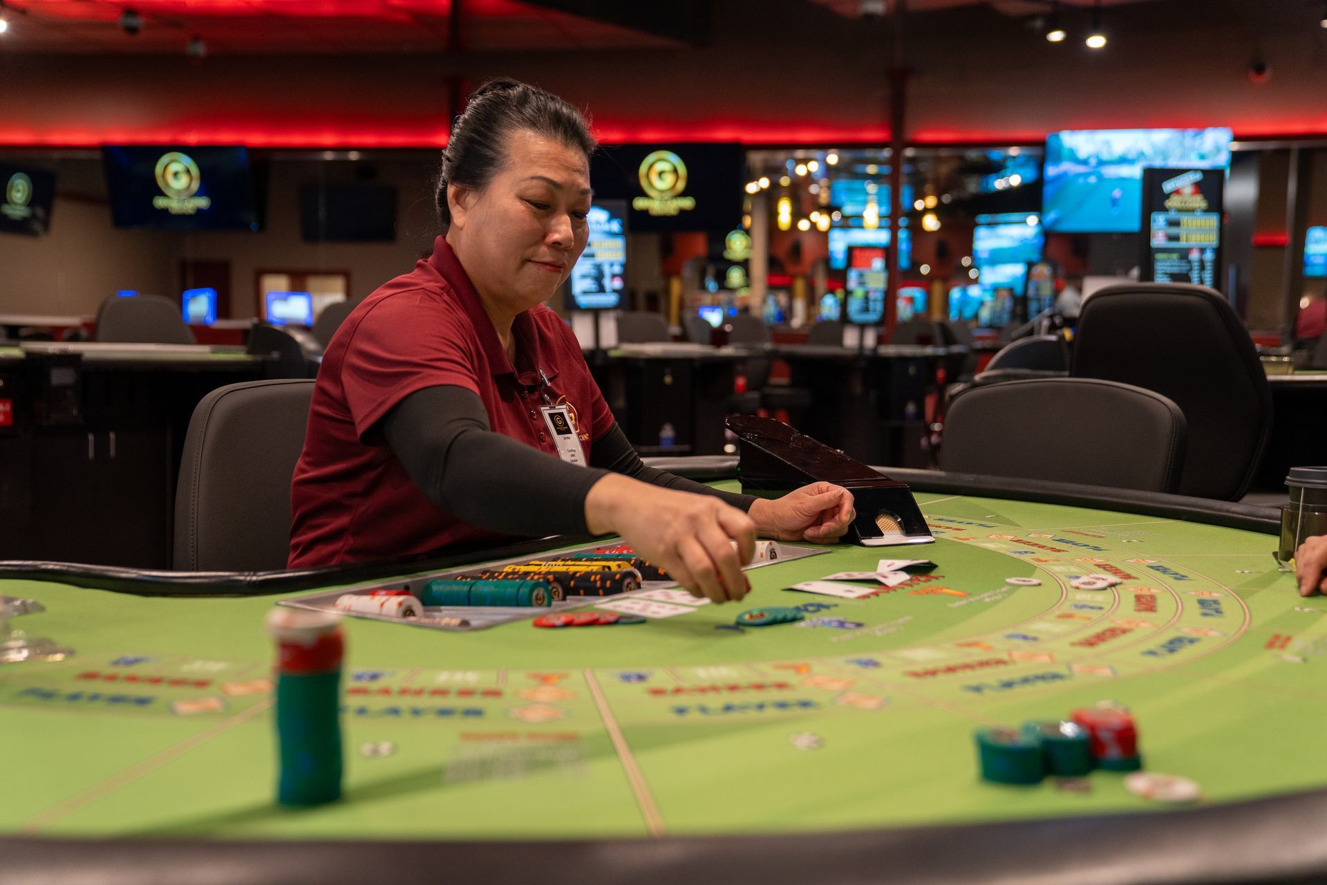 Empty casino floor featuring round gaming tables with chairs on patterned dark carpet under warm, geometric lighting.