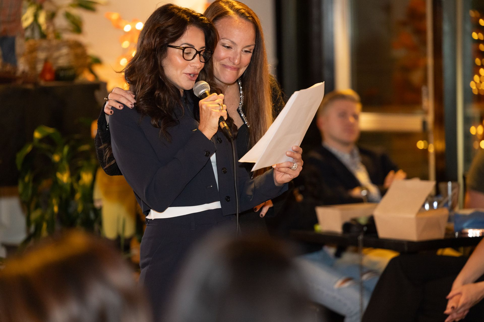 Two women are standing next to each other holding a microphone and reading a piece of paper.