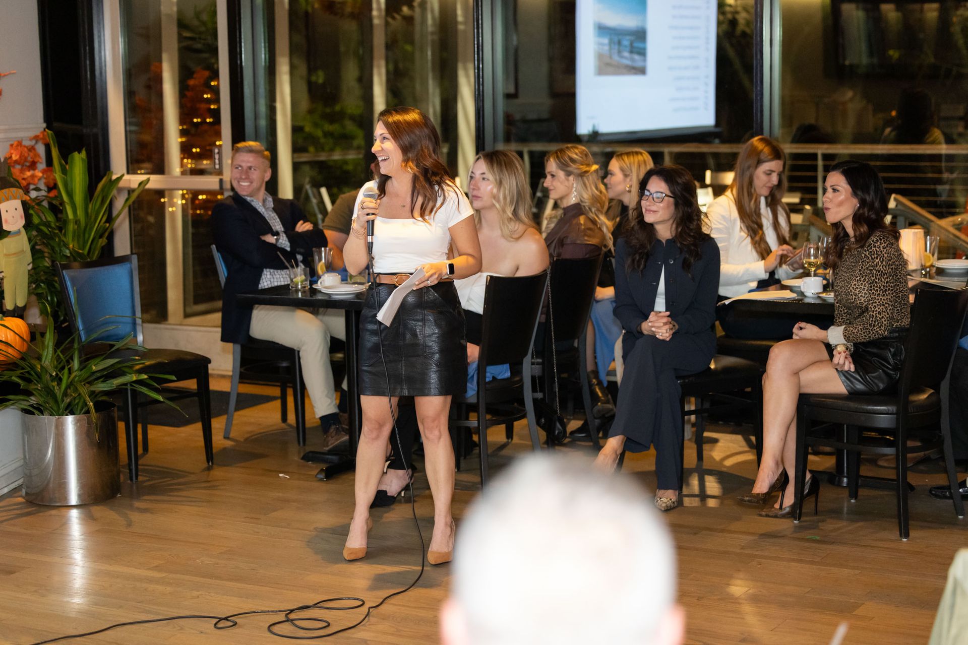 A woman is standing in front of a microphone while a group of people sit at tables.