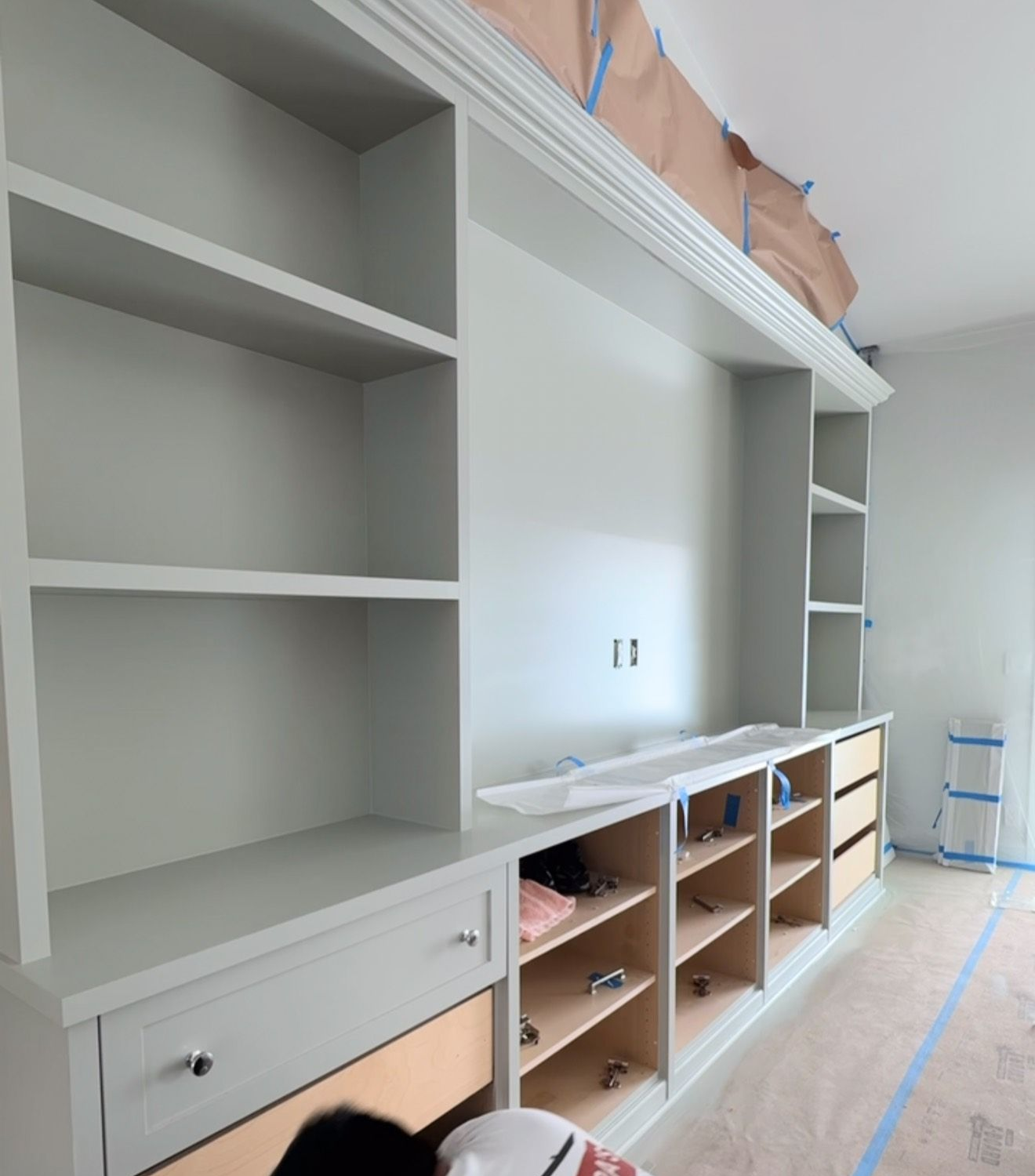 Built-in white shelving and cabinets in a room under renovation, with open cubbies and taped floor coverings